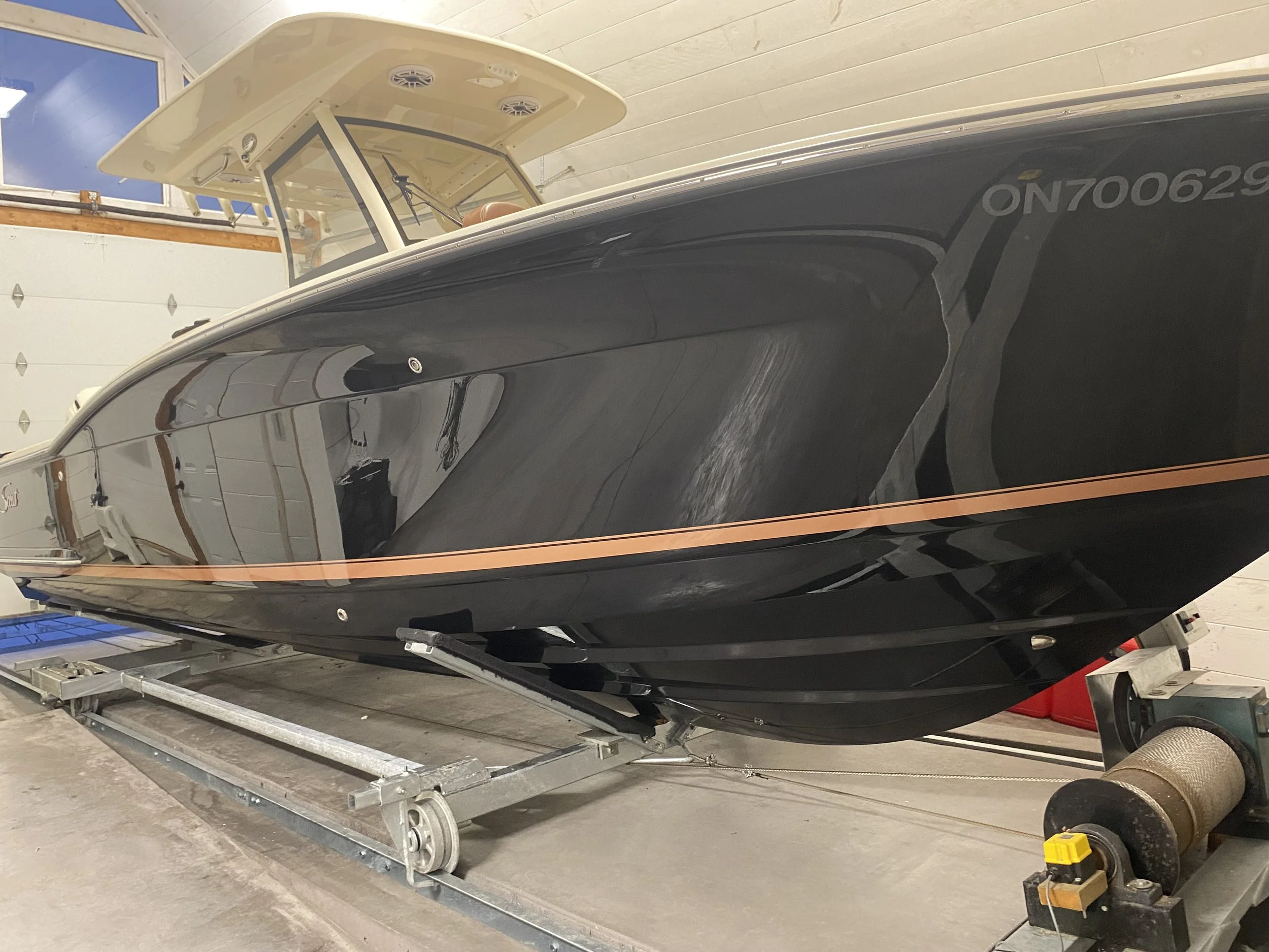 A black boat with a tan stripe, stored indoors on a trailer, with windows and a white top section.