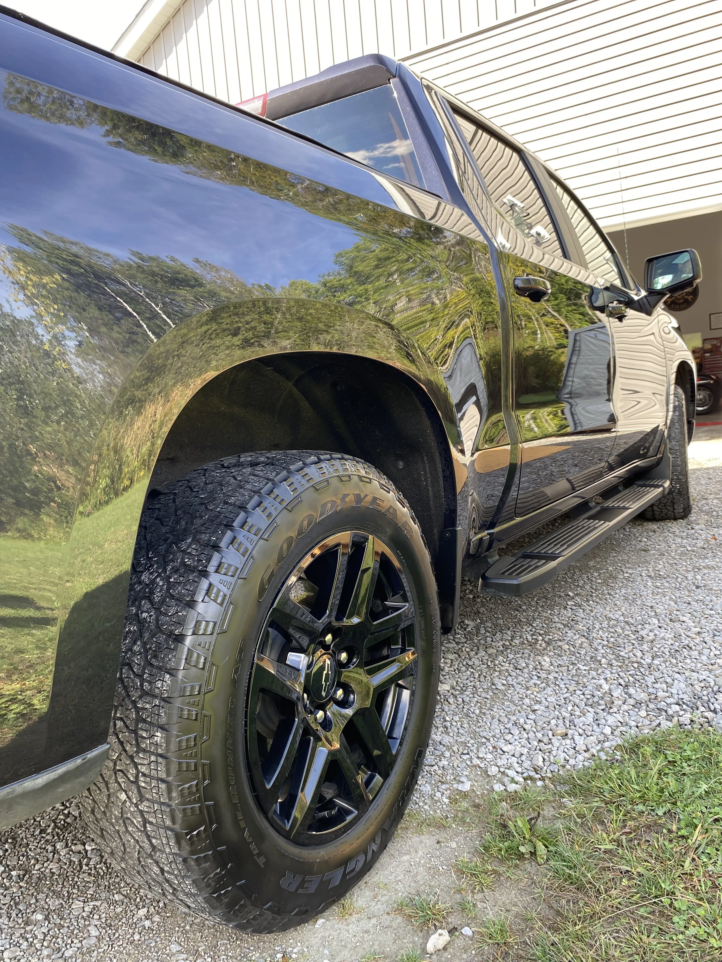 A black pickup truck parked on a gravel driveway in front of a white building, with reflections of trees visible on the truck's surface.