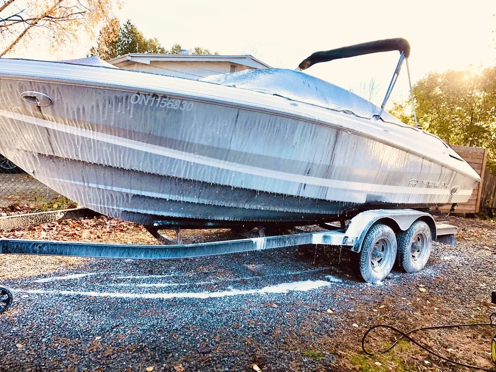 A boat on a trailer being washed with a hose, with water and soap runoff on the ground.