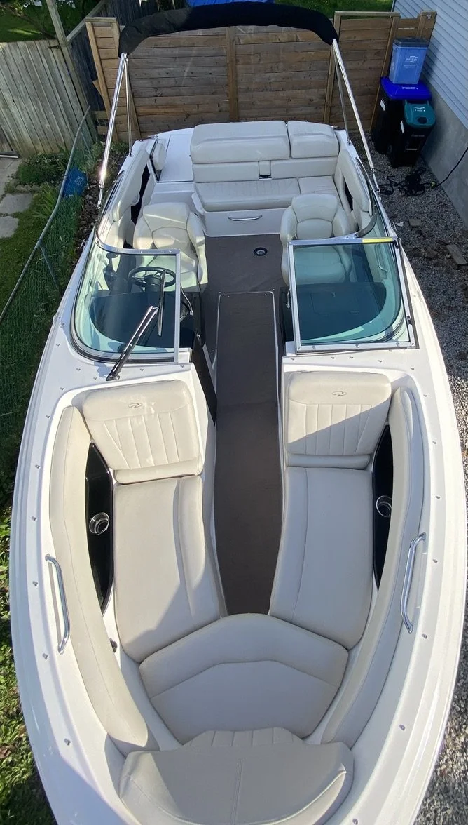 A white motorboat with beige cushioned seats, viewed from above, parked near a wooden fence and a house, with a utility area containing trash bins in the background.