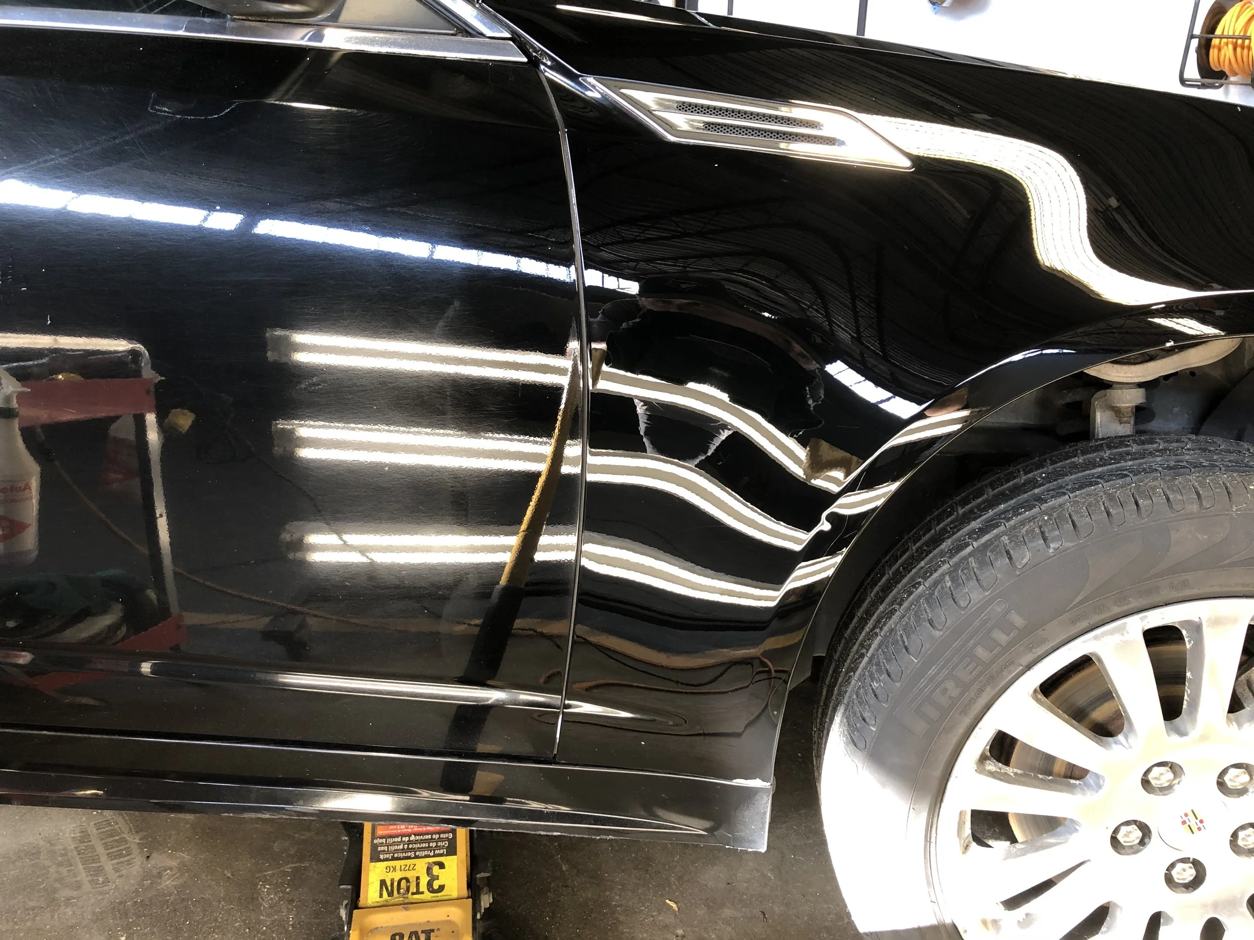 Close-up of a black car's front passenger side with wheel, reflecting fluorescent lights and workshop environment.