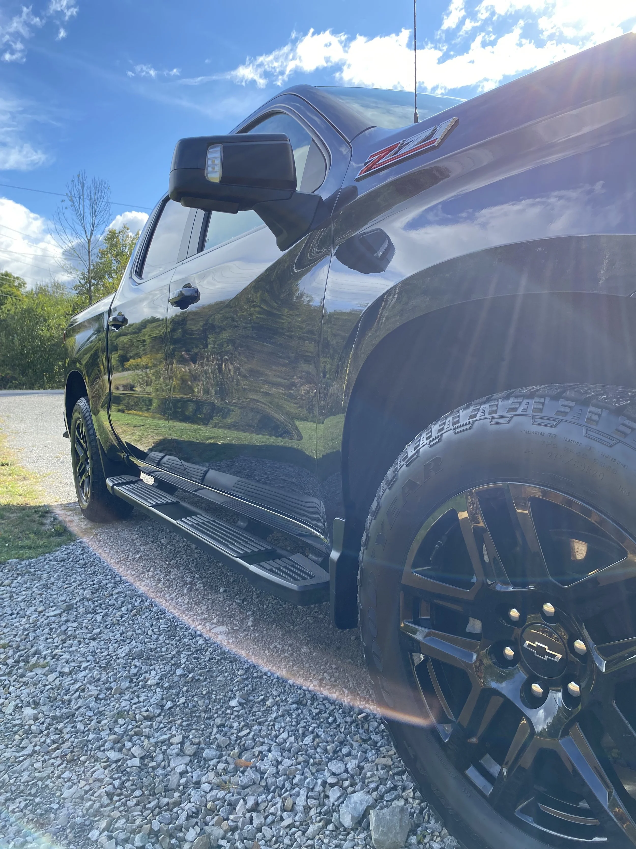 Black Chevrolet pickup truck parked on gravel with a blue sky and trees in the background, sunlight creating lens flare.