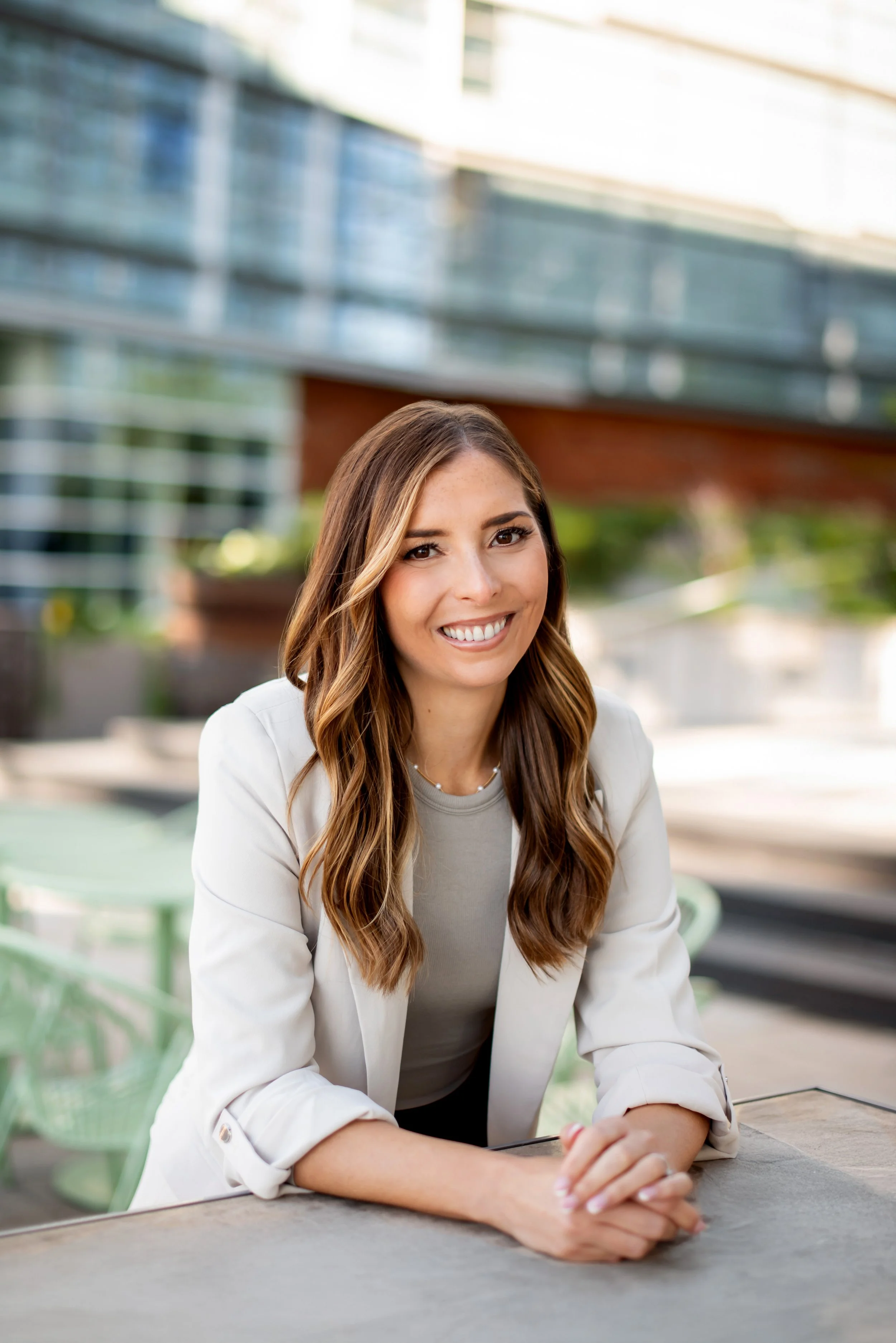 A young woman with long wavy brown hair, wearing a light blazer, is smiling and sitting outside at a table in an urban area with modern buildings in the background.