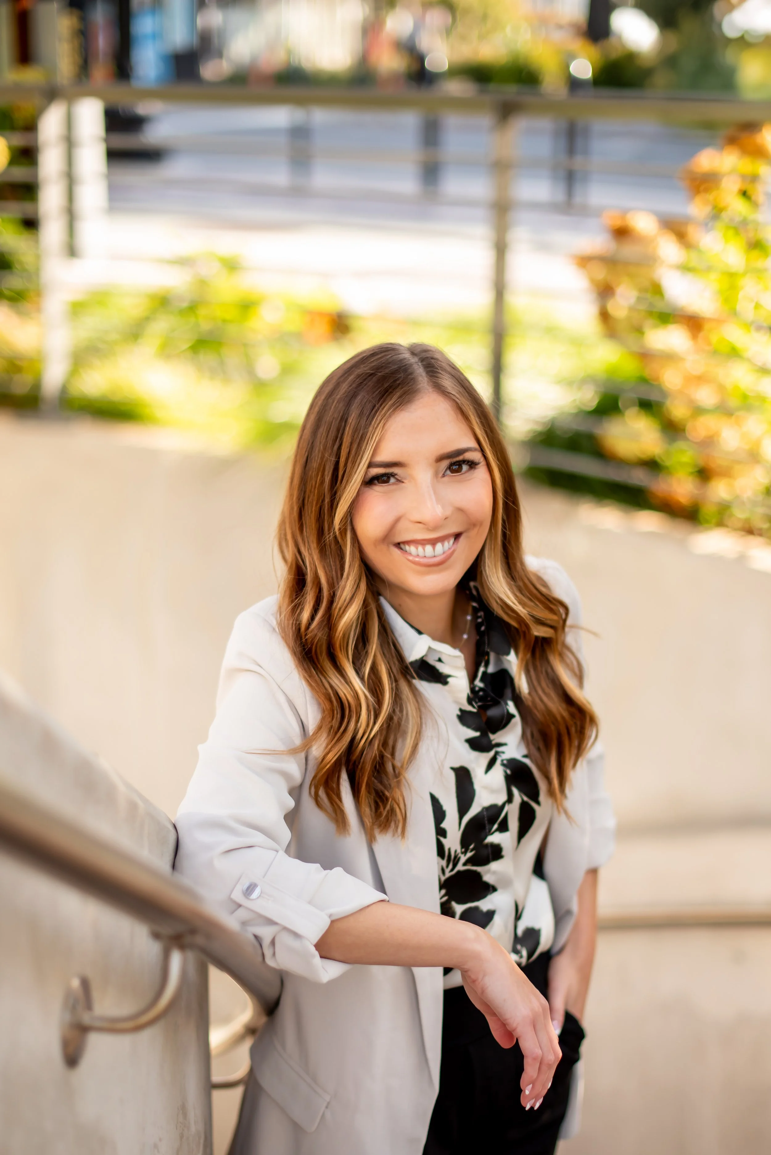 A young woman with long, wavy brown hair and a white blazer, smiling and leaning on a railing outdoors during daytime.