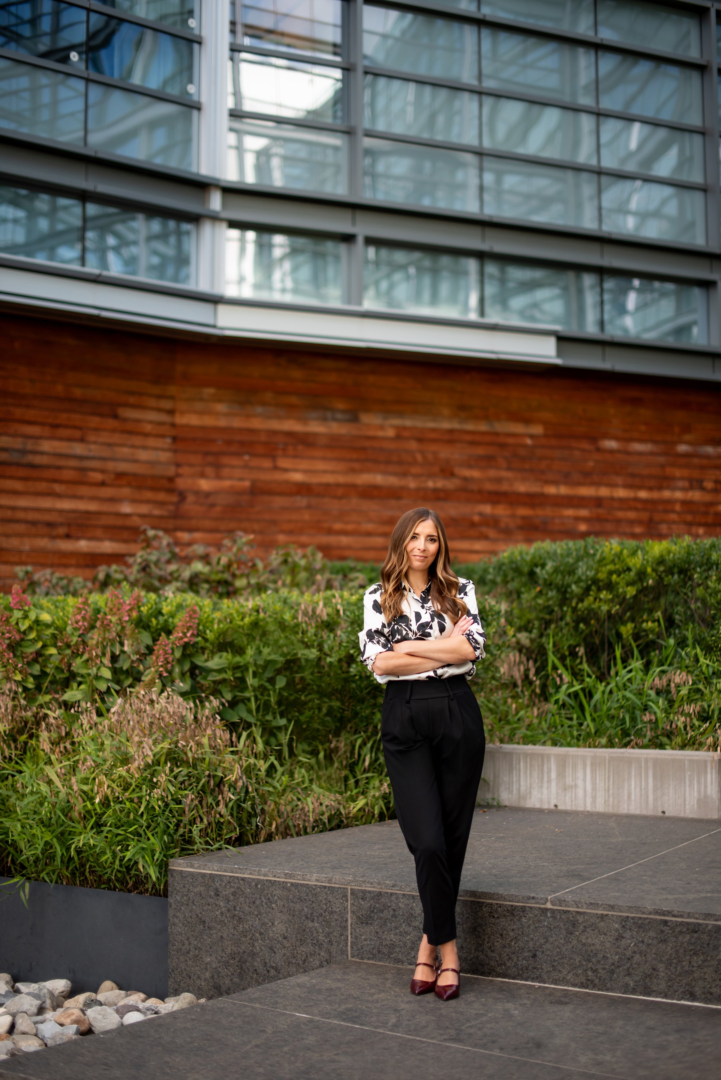 A woman with long wavy brown hair wearing a black and white patterned blouse, black high-waisted pants, and burgundy high heels standing in front of greenery and a modern building with glass windows and a wooden panel wall.