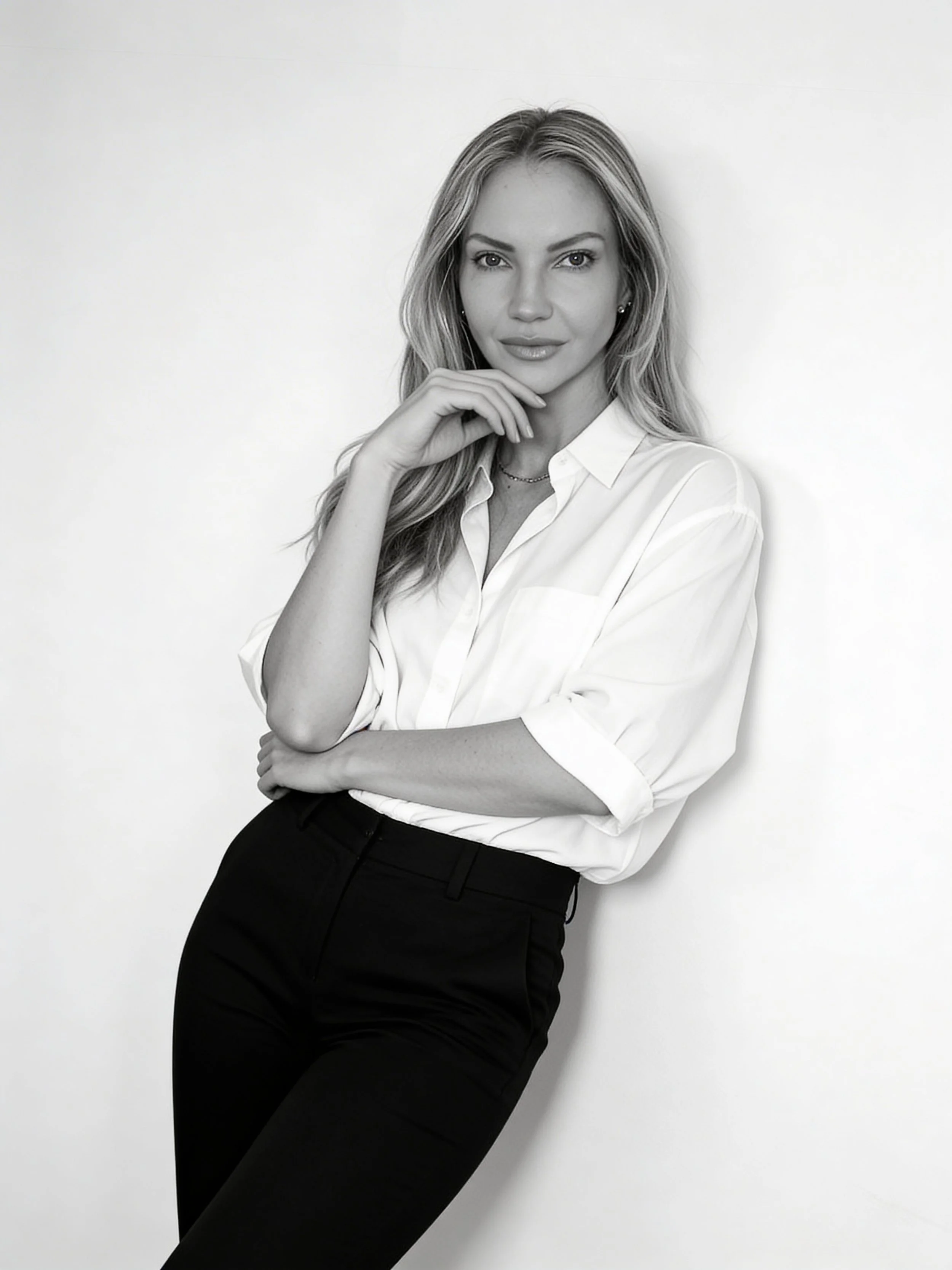 Black and white photo of a young woman with long wavy hair, wearing a white blouse and black pants, standing against a plain white wall, with one hand near her chin and a confident expression.