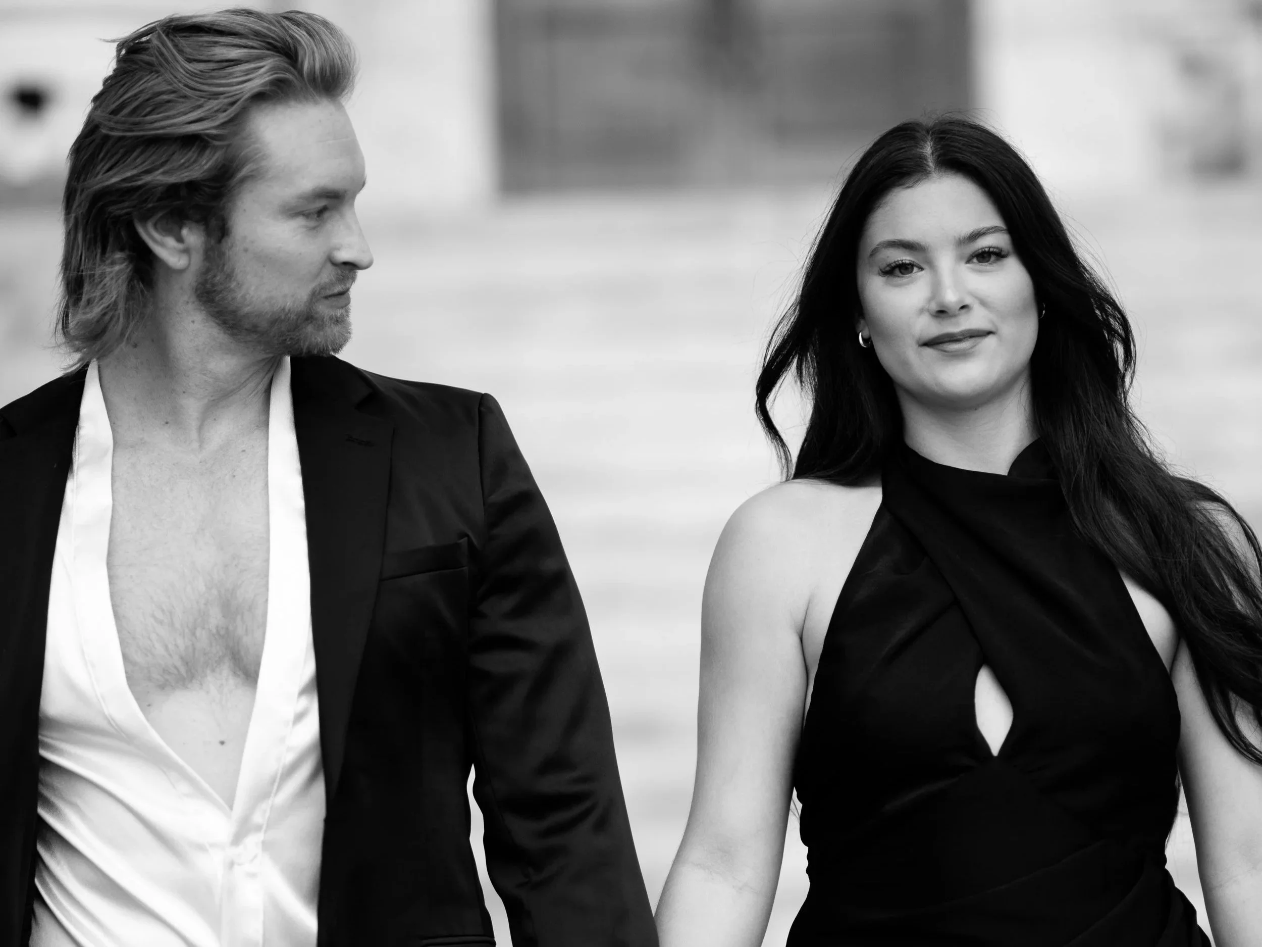 A black and white photo of a man and a woman standing close together, looking at each other. The man has shoulder-length hair and a beard, wearing a blazer over a partially unbuttoned shirt. The woman has long, dark hair, wears earrings, and is dressed in a sleeveless top.