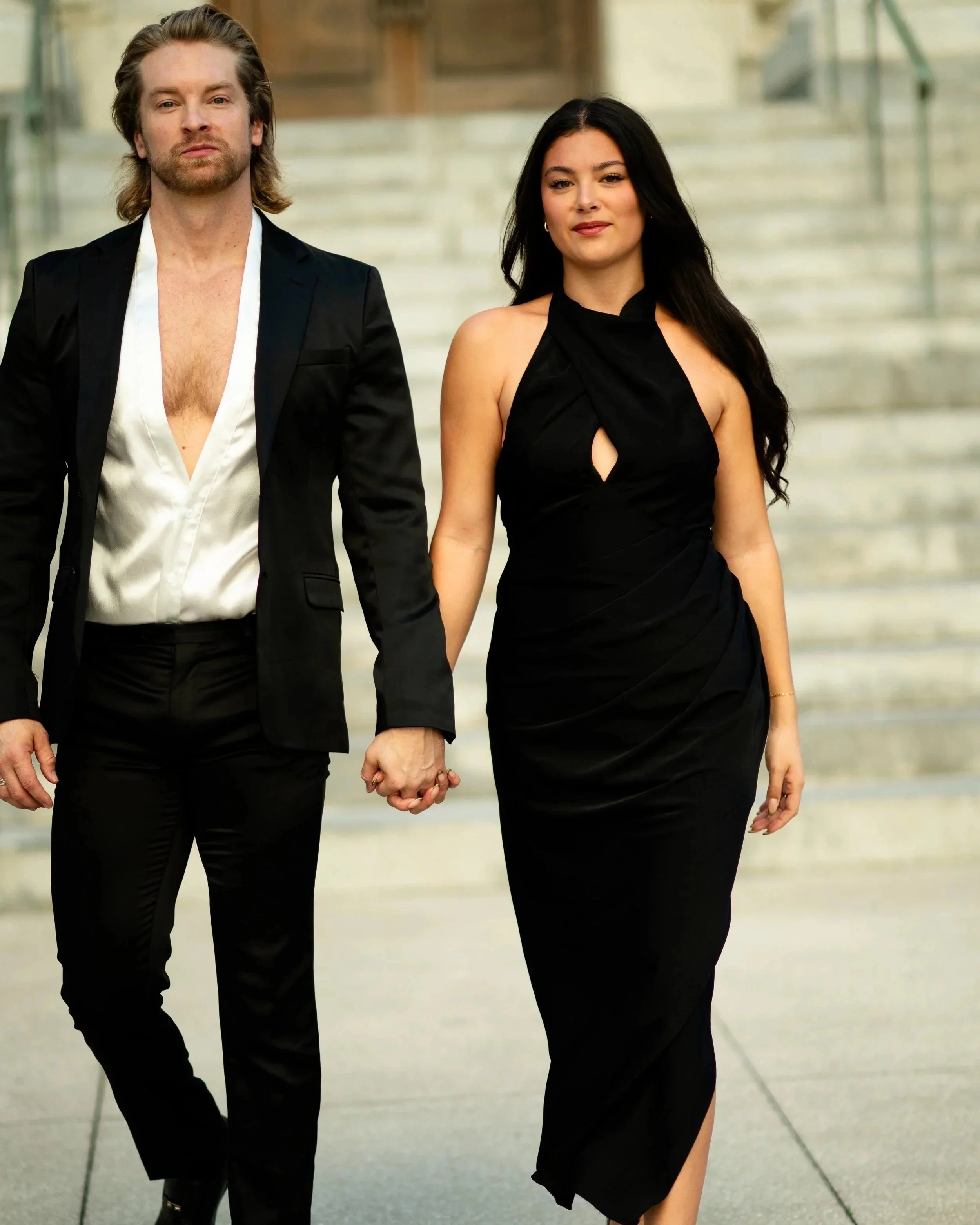A man and woman holding hands walking outdoors. The man wears a black blazer, open white shirt, and black pants, and has shoulder-length hair and a beard. The woman wears a black dress with a slit, has long dark hair, and is smiling slightly. In the background, there are stairs and a glass railing.