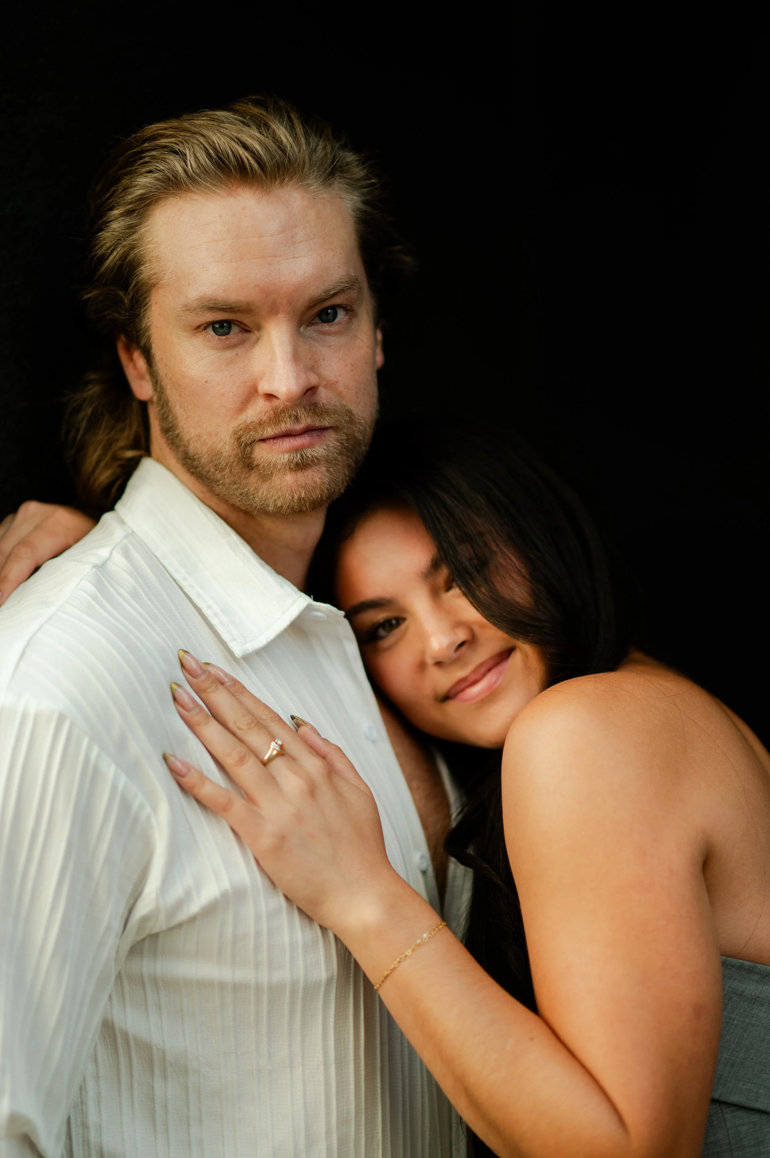 A man with blonde hair and a beard wearing a white shirt, and a woman with dark hair and a warm smile hugging him, against a black background.