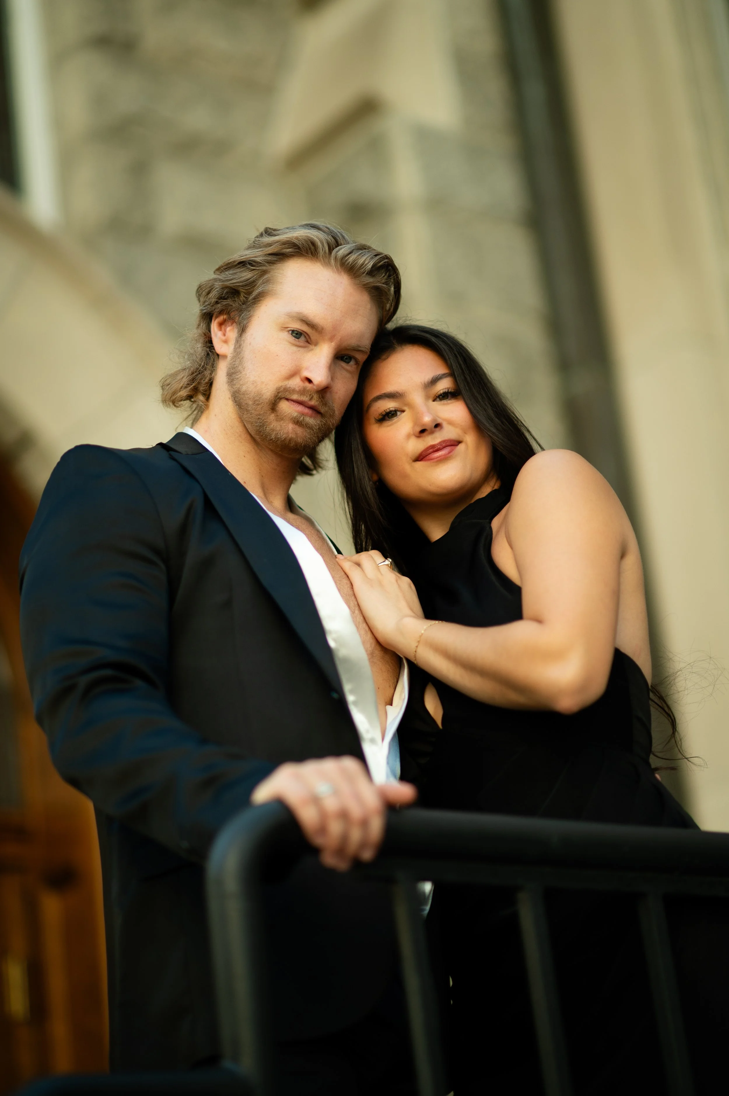 A man and woman posing closely in formal attire against a blurred background, possibly outdoors or near a building.