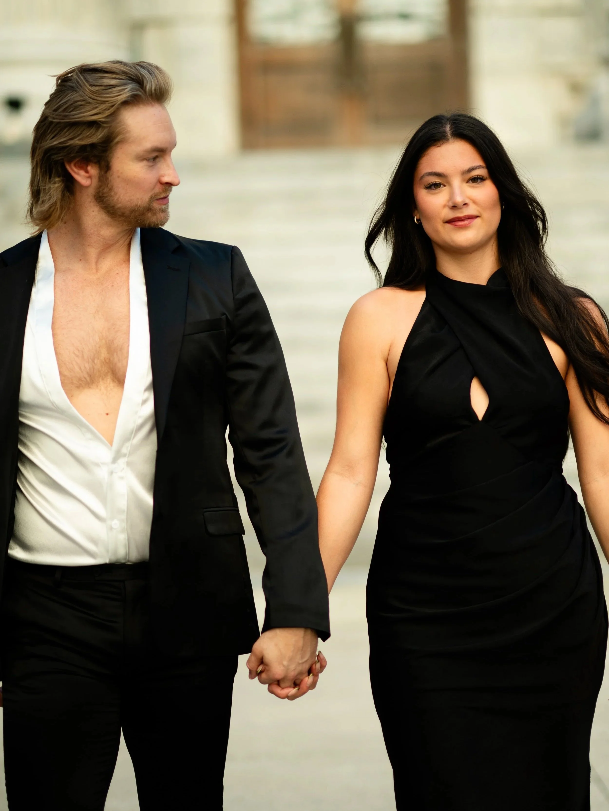 A man and woman holding hands, walking outdoors in formal attire, with the man wearing a partially unbuttoned shirt and the woman in a black dress.