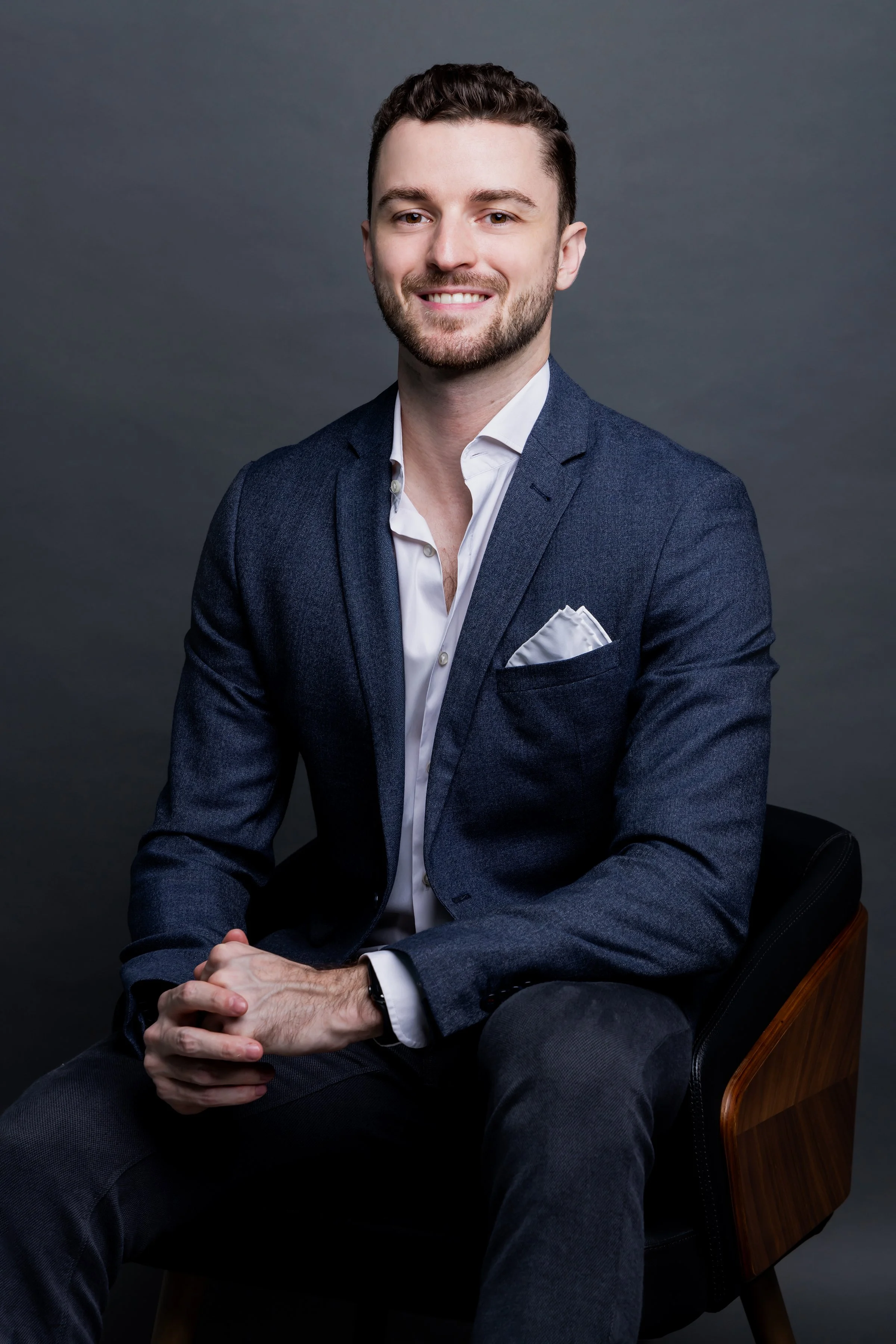 A young man in a dark blue suit and white shirt sitting on a black and wooden chair against a plain gray background.