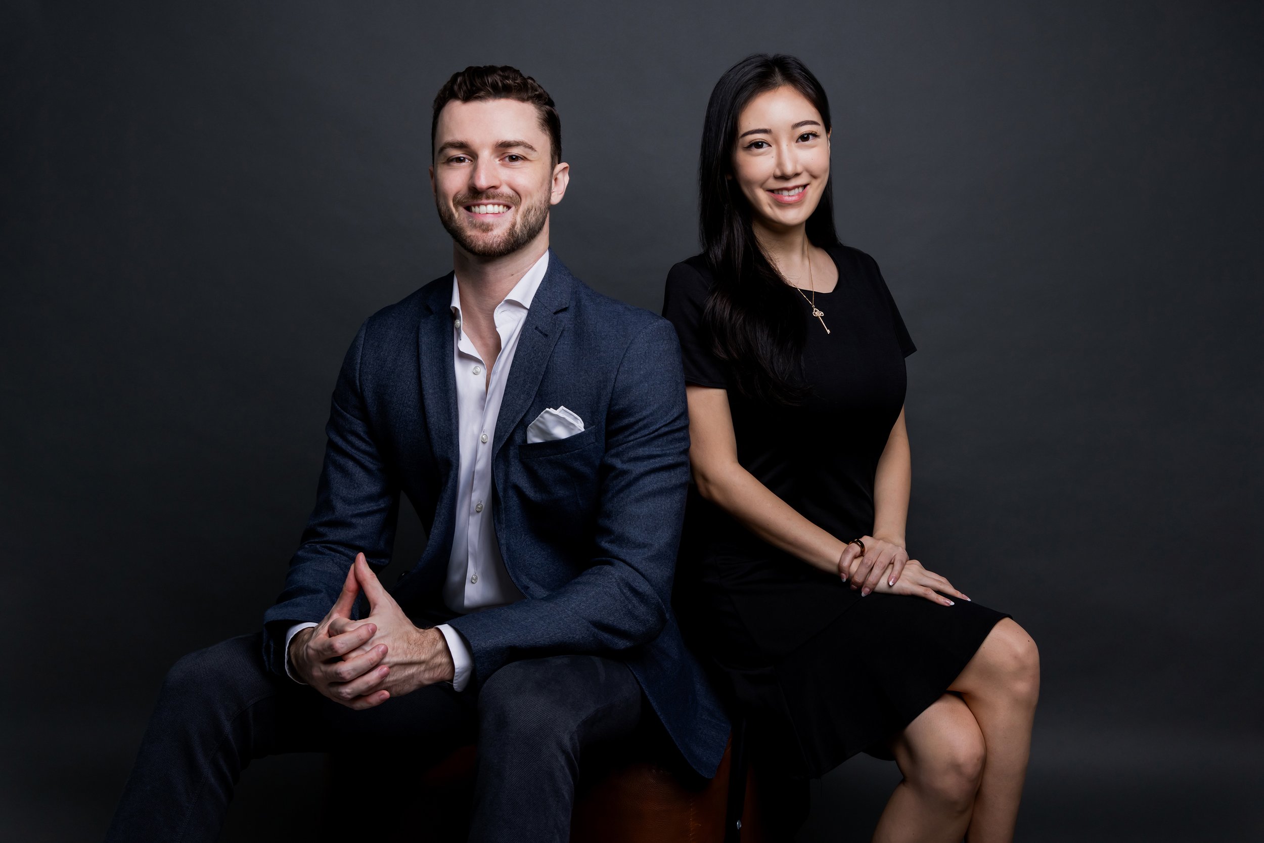 A man and a woman sitting against a dark gray background, smiling at the camera. The man is wearing a navy business suit with a white shirt and pocket square, and the woman is dressed in a black dress with a necklace.