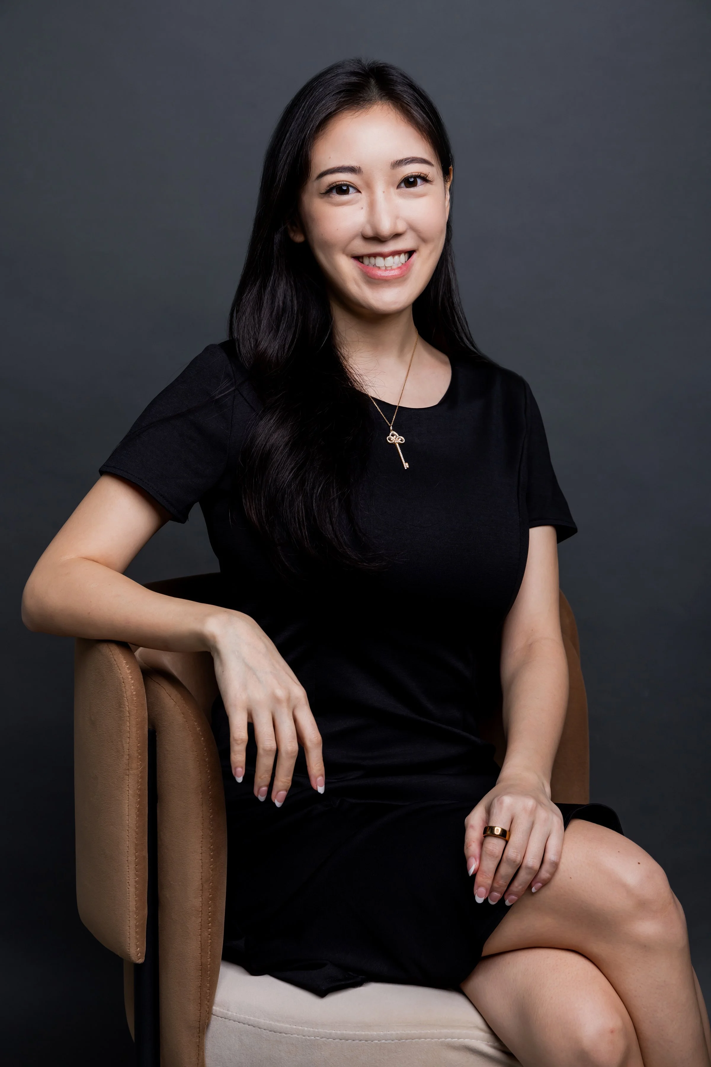 A young woman with long dark hair, wearing a black dress and a key necklace, smiling while sitting on a beige chair against a dark grey background.