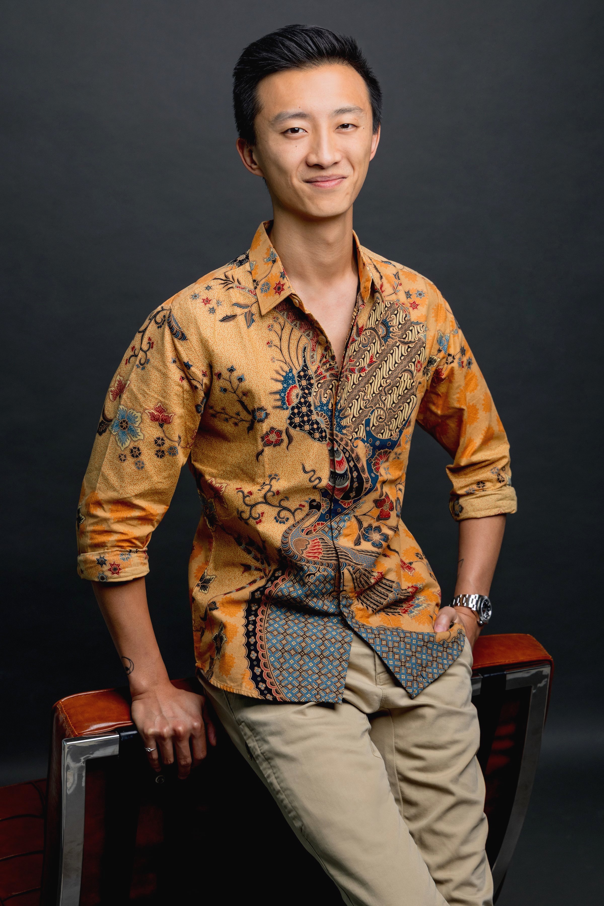 A young Asian man with dark hair, wearing a patterned orange and brown shirt and beige pants, sitting on a stool against a dark background.
