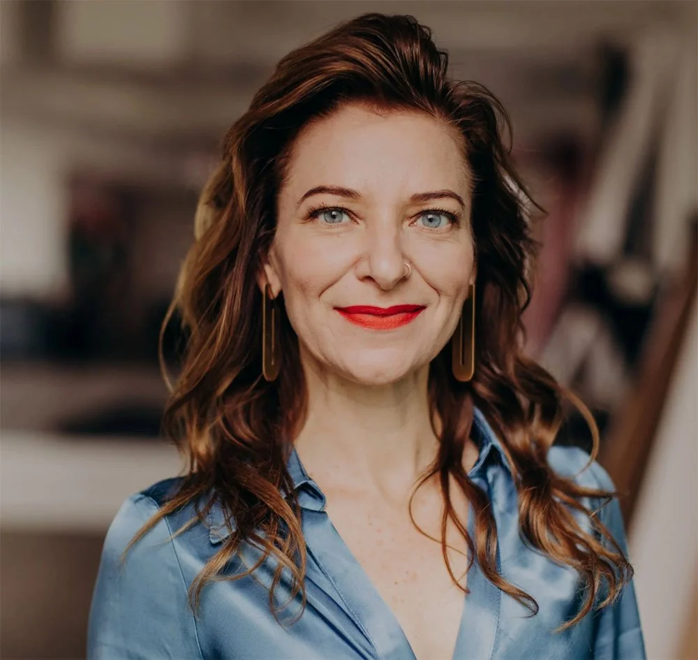 Portrait of a woman with red hair, wearing a coral headband, green top, and denim jumper, standing against a beige wall outdoors.