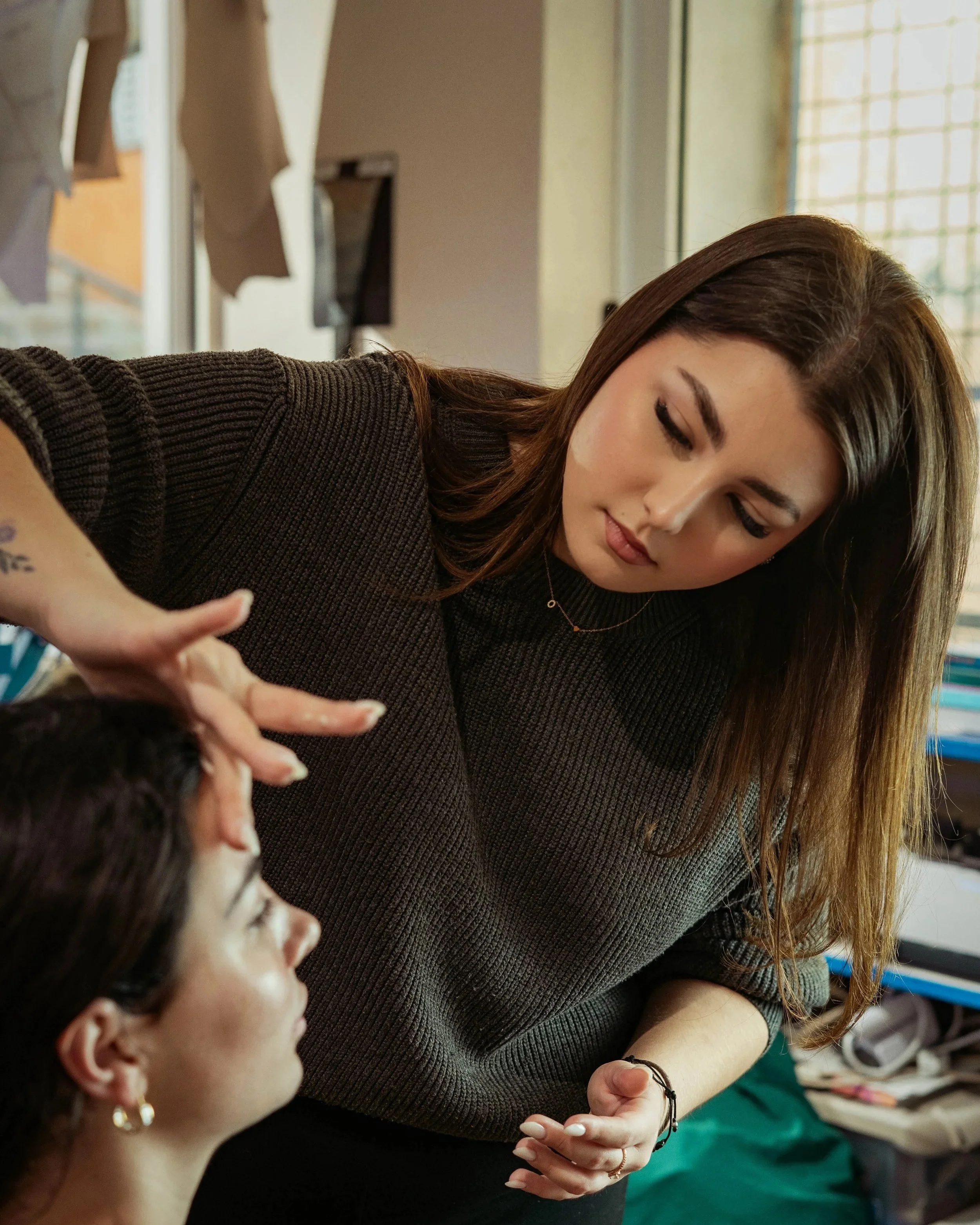 Una mujer con cabello castaño y trenzado, vestida con un suéter marrón oscuro, atiende a otra mujer en un entorno interior, posiblemente una peluquería o salón de belleza, mientras la segunda mujer tiene el cabello recogido y lleva pendientes dorados.
