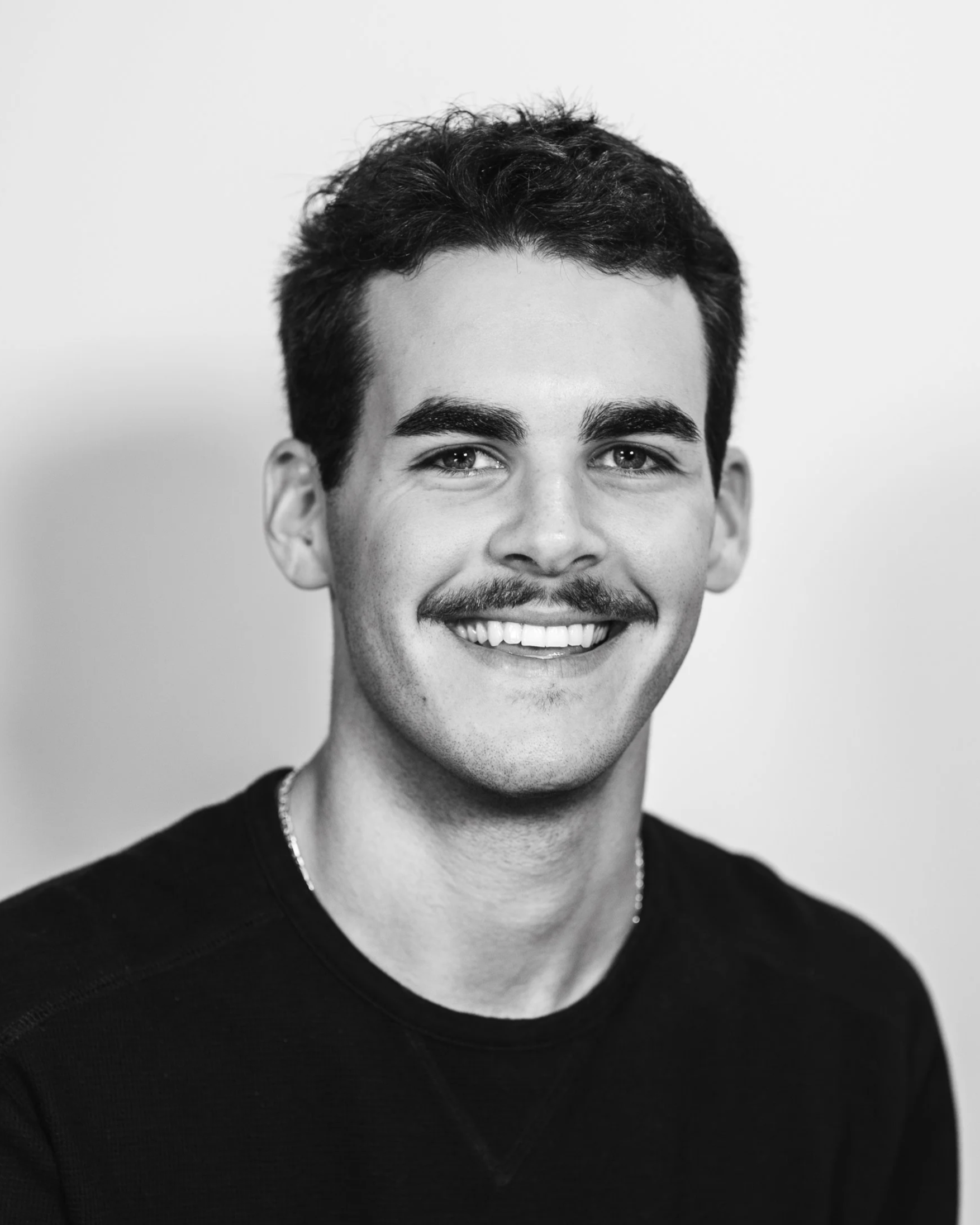 Black and white portrait of a smiling young man with dark hair, thick eyebrows, a mustache, wearing a black shirt.