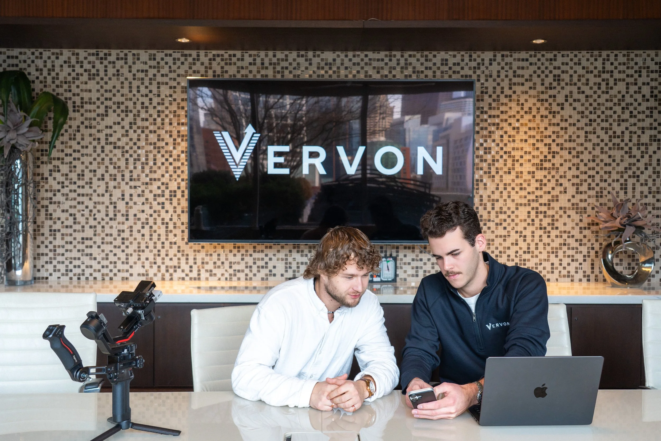 Two men seated at a conference table, one in a white shirt and the other in a dark jacket, working with a laptop and smartphone in front of a large screen displaying the logo "Veron".