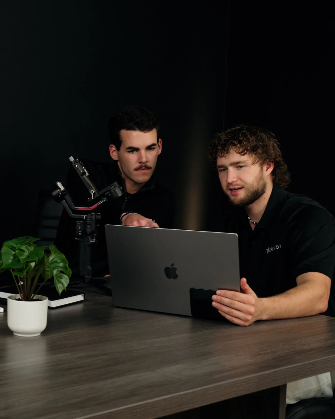 Two men working together at a desk with a laptop, a potted plant, and a notebook, in a black room.