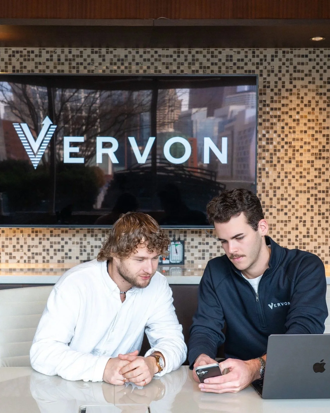 Two men sitting at a table, one wearing a white shirt and the other in a navy jacket with VERVON logo, looking at a smartphone, with a laptop open on the table. Behind them is a large screen displaying the VERVON logo.