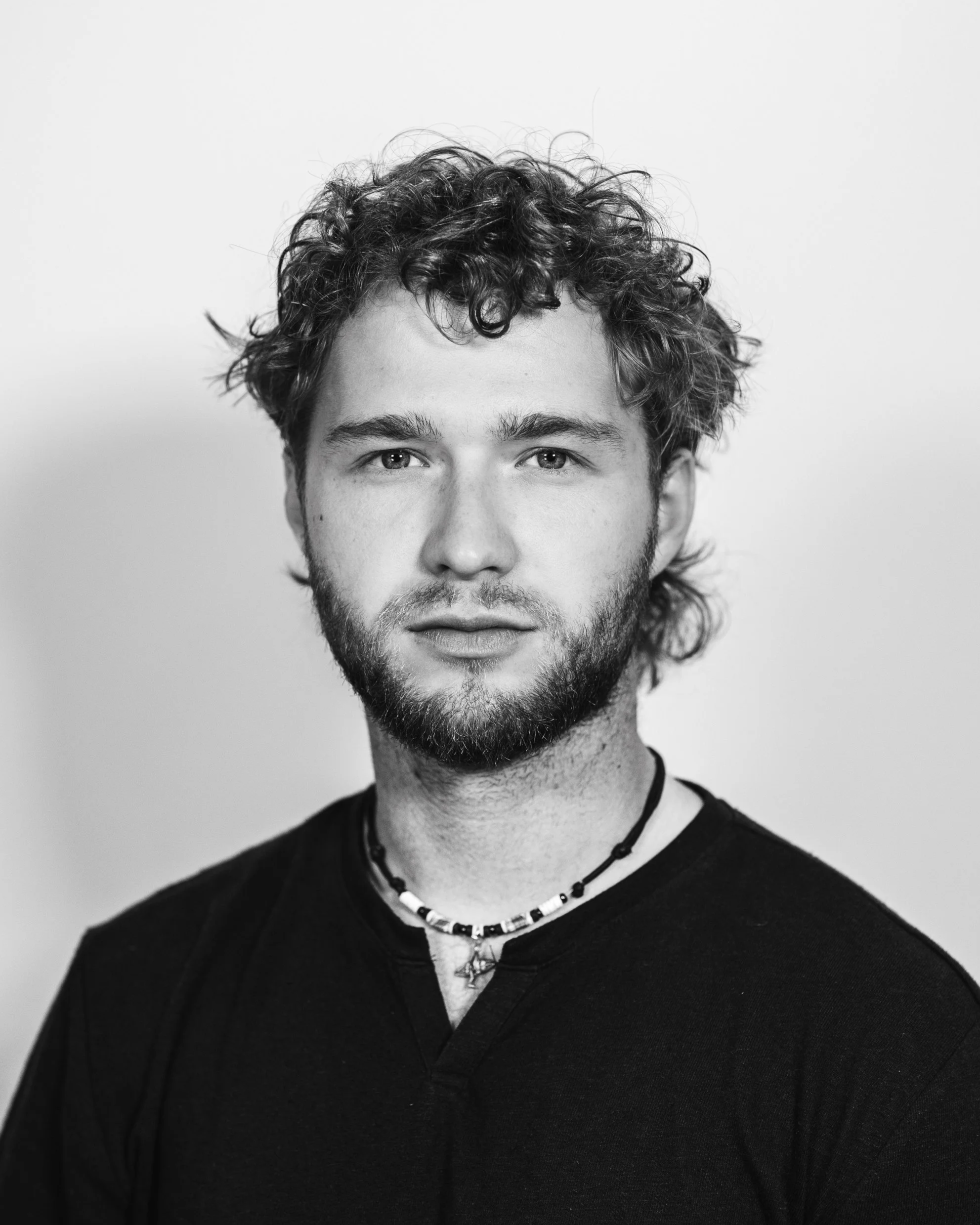 Black and white portrait of a young man with curly hair and a beard, wearing a dark shirt and a beaded necklace, looking directly at the camera.