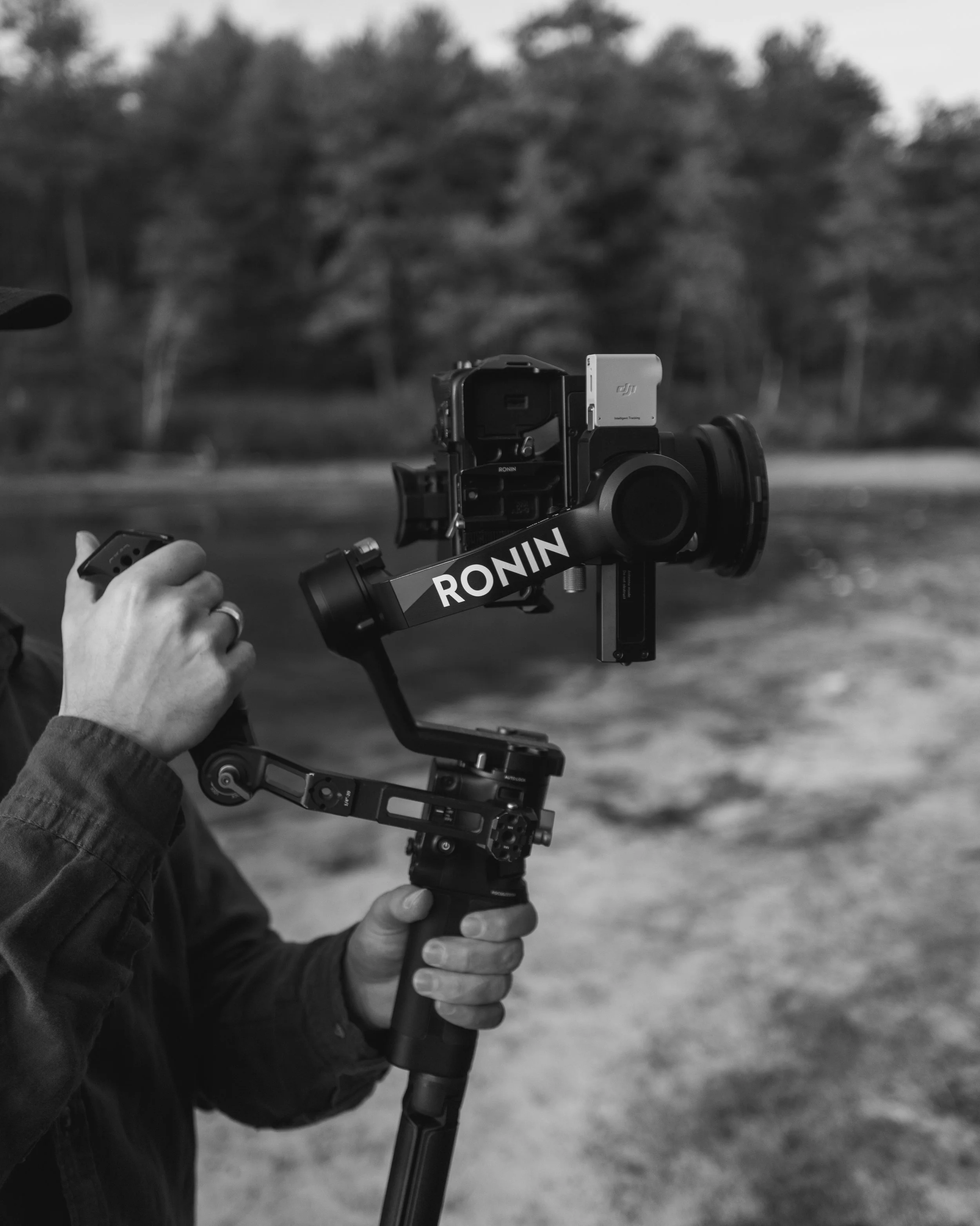 A person operating a camera mounted on a handheld stabilizer with the brand name RONIN printed on it, set outdoors with trees blurred in the background.