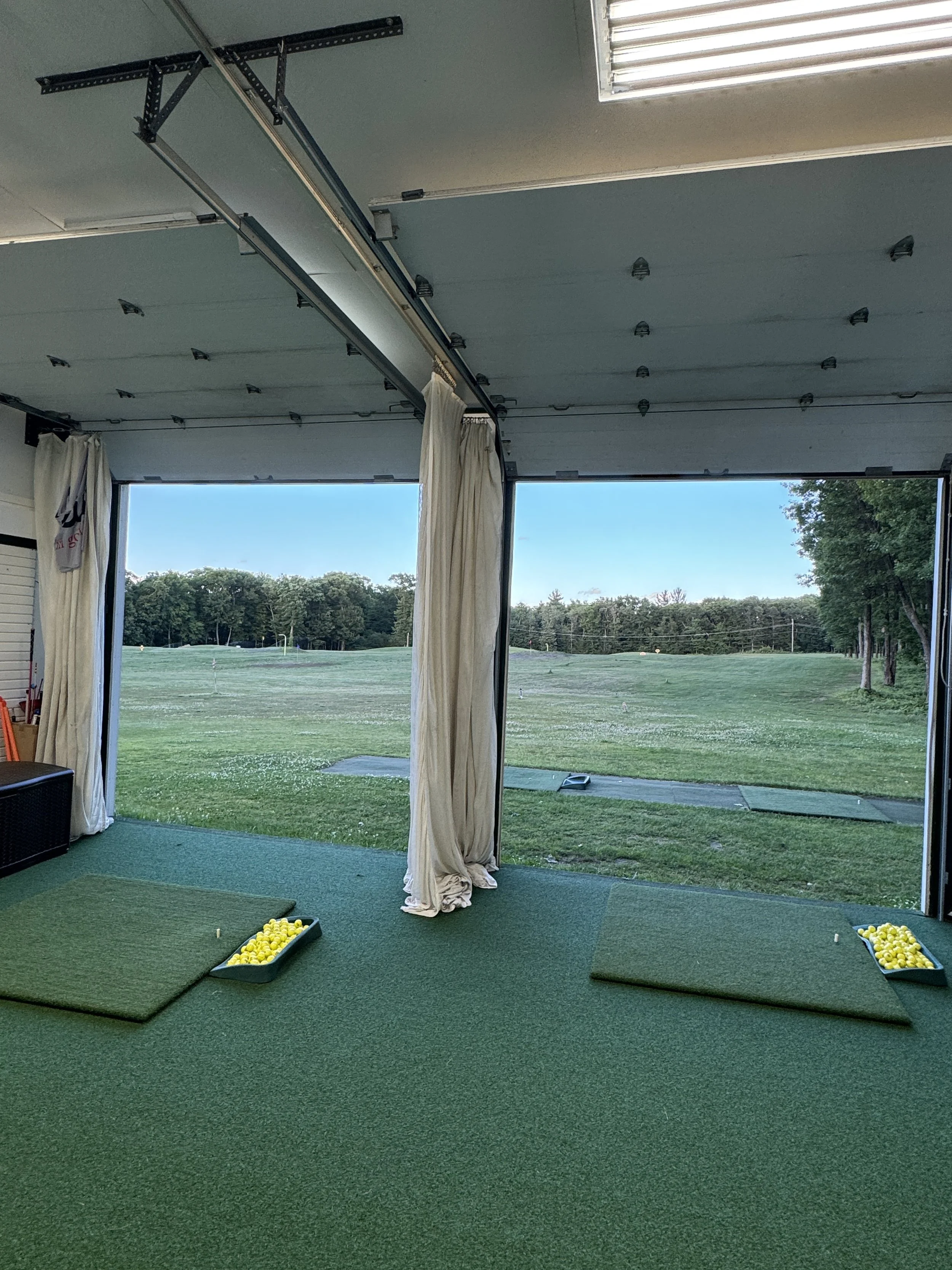 Indoor golf practice area with artificial turf, opening to outdoor golf range, with yellow golf balls in trays, white driving range nets, and an open large garage door.  This is where golf lessons are given year-round during all 4 seasons.