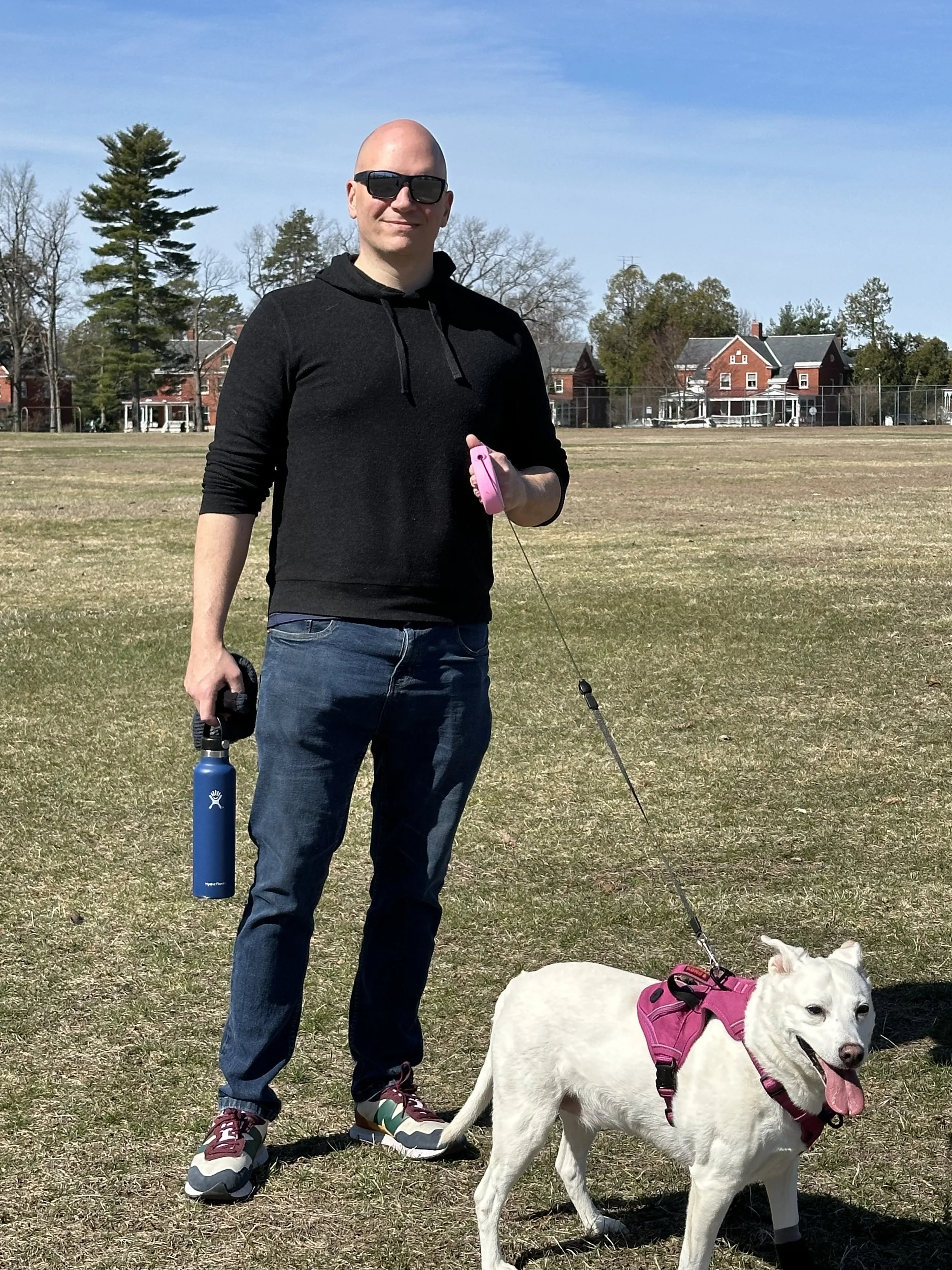 A man with sunglasses standing outdoors on a grassy field with his white dog, both on leashes. The man wears a black hoodie, jeans, and sneakers. The dog has a pink harness and is panting.