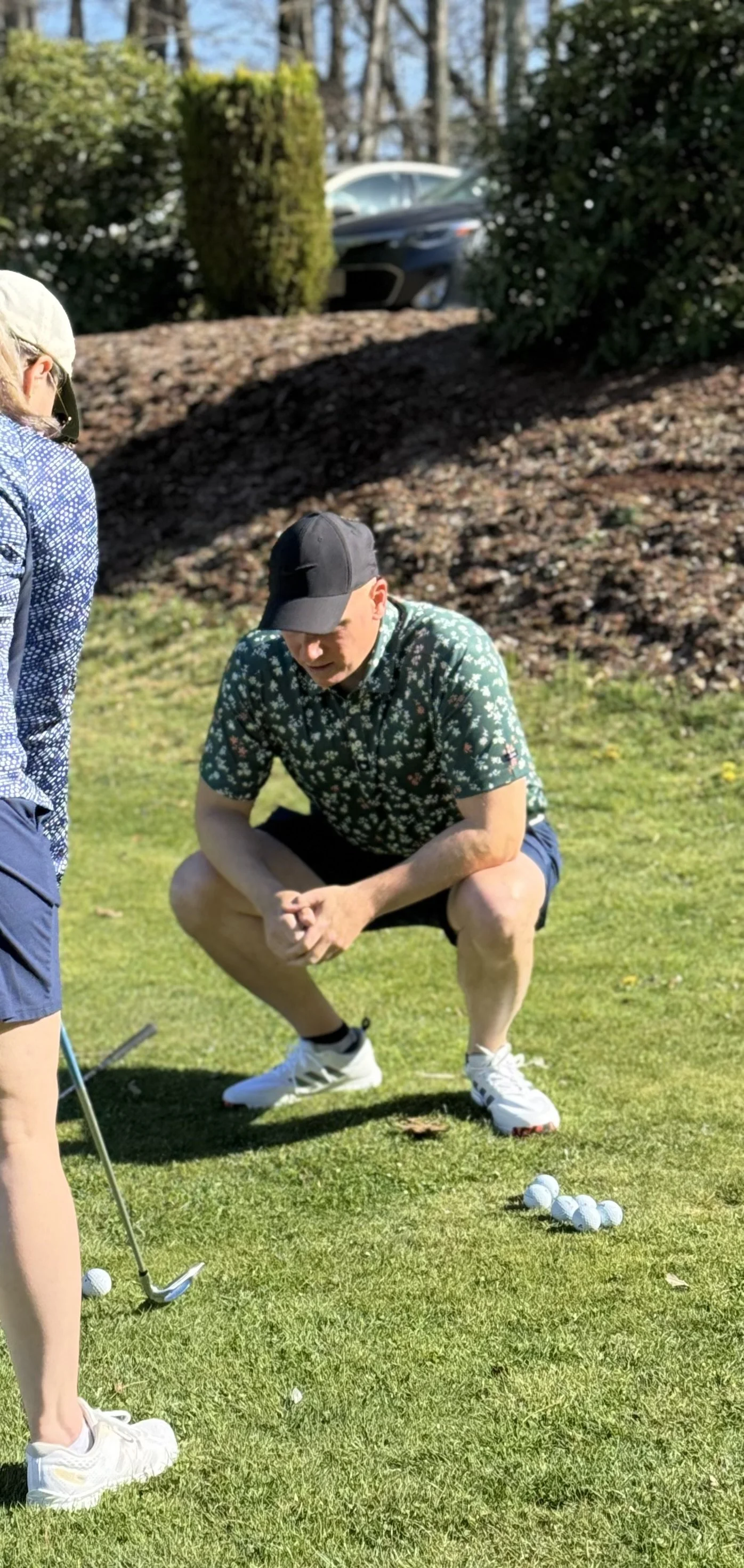 A golf instructor crouching on grass next to a woman golf student who is taking a golf lesson with golf clubs and golf balls during a sunny day.