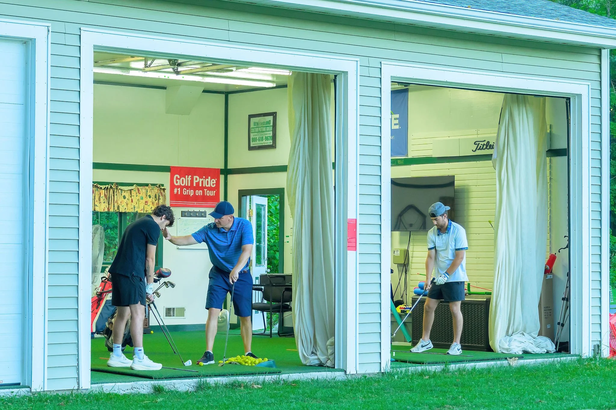 One man, a golf instructor, and a boy practicing hitting golf balls from indoors under the roof out onto the outdoor driving range, with the golf instructor helping the boy with his golf swing, and another person practicing their own golf shot.