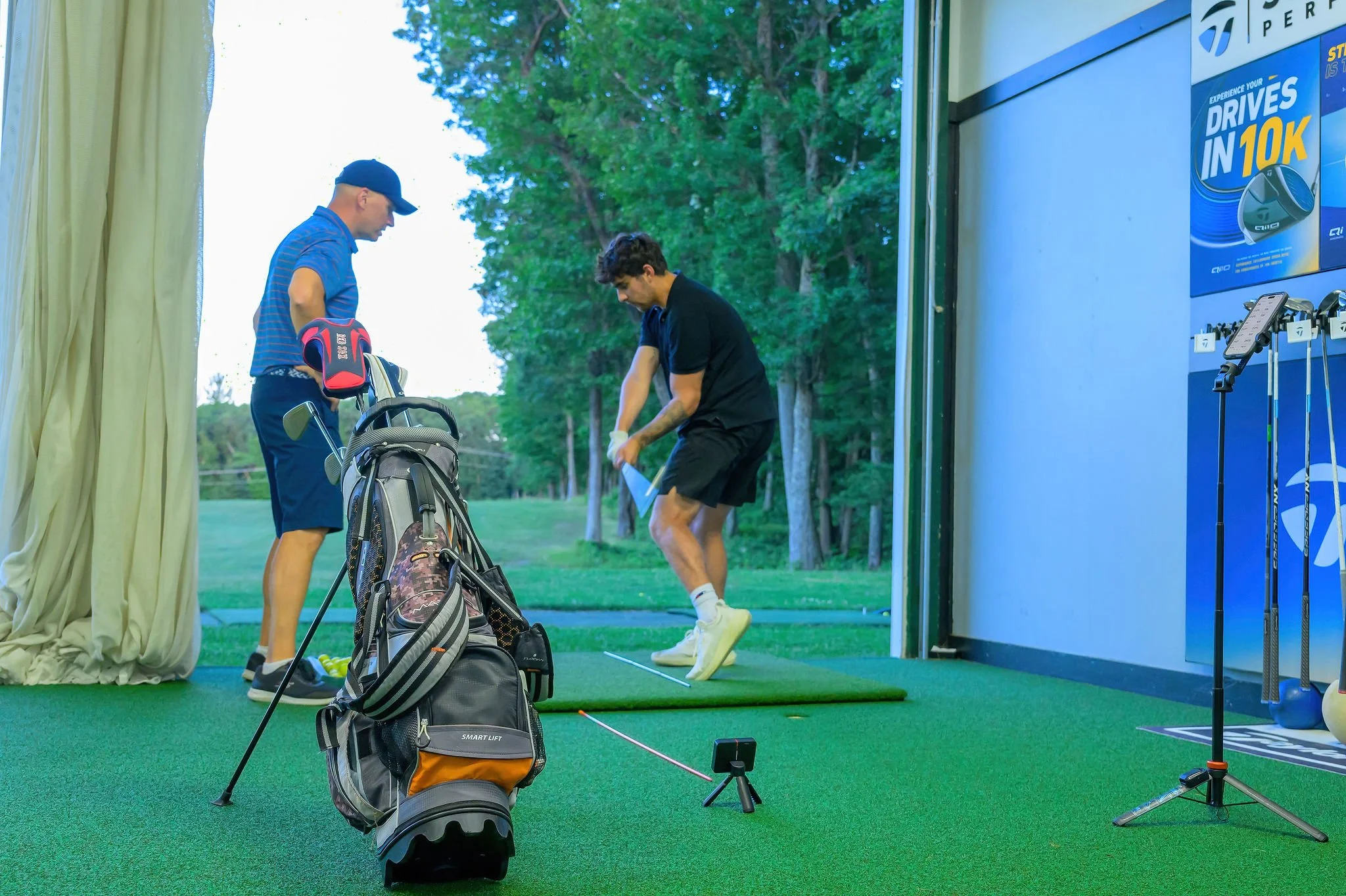 One man and a golf instructor practicing golf indoors; one is swinging a club on a green driving range mat, the golf instructor is observing with a golf club in hand. Golf clubs, a golf bag, and a small golf ball launch monitor are nearby.