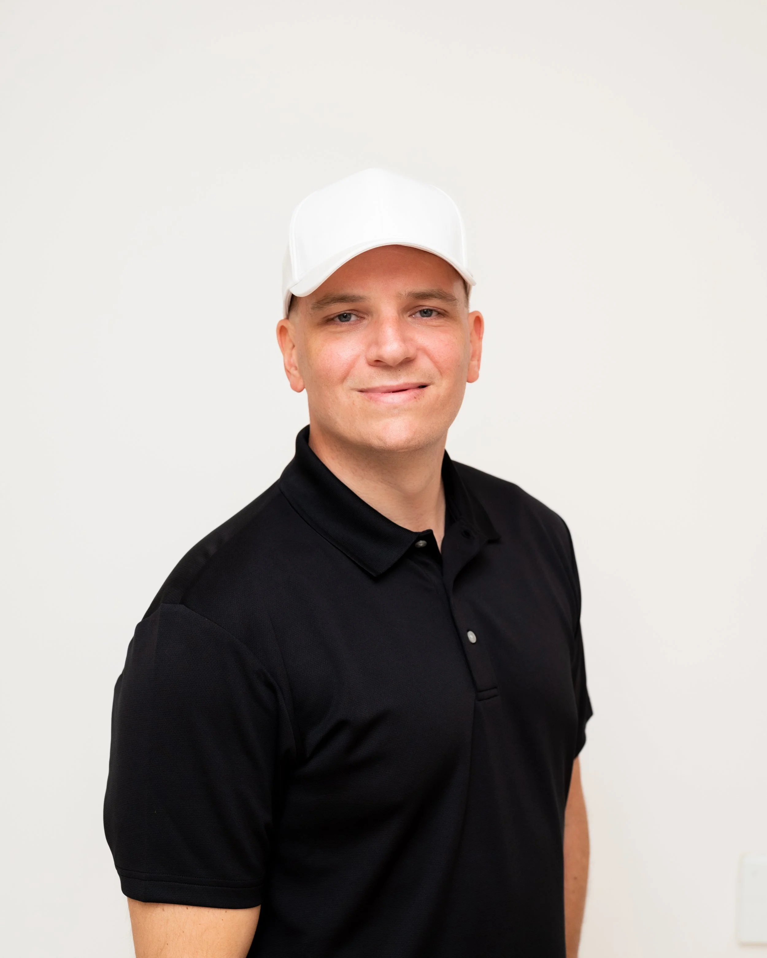 Andy Merrill, the man wearing a black polo shirt and a white cap, standing against a white wall, smiling at the camera, is the head golf instructor for 495 Golf which is a golf instruction business serving eastern and central Massachusetts.