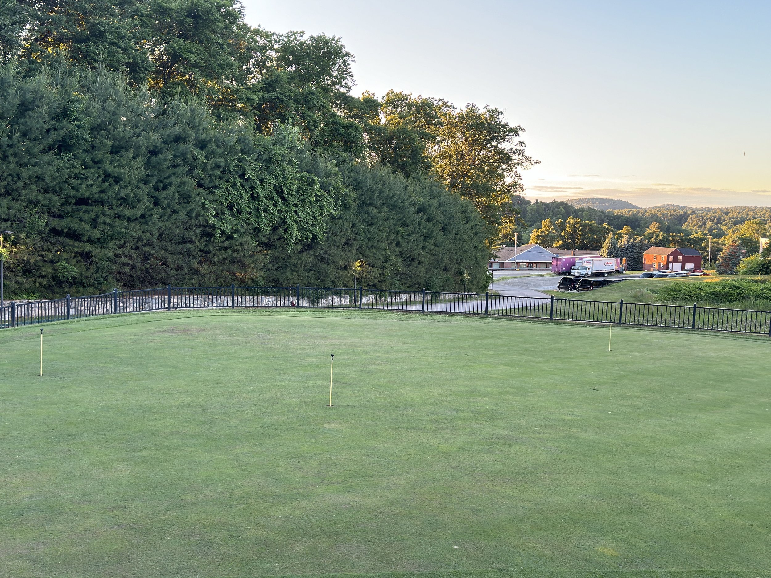 Golf course green with three flagsticks, surrounded by a black metal fence, with a wooded hill on the left and this is where short game golf lessons are given that focus on the golf skills of putting, chipping, and pitching.