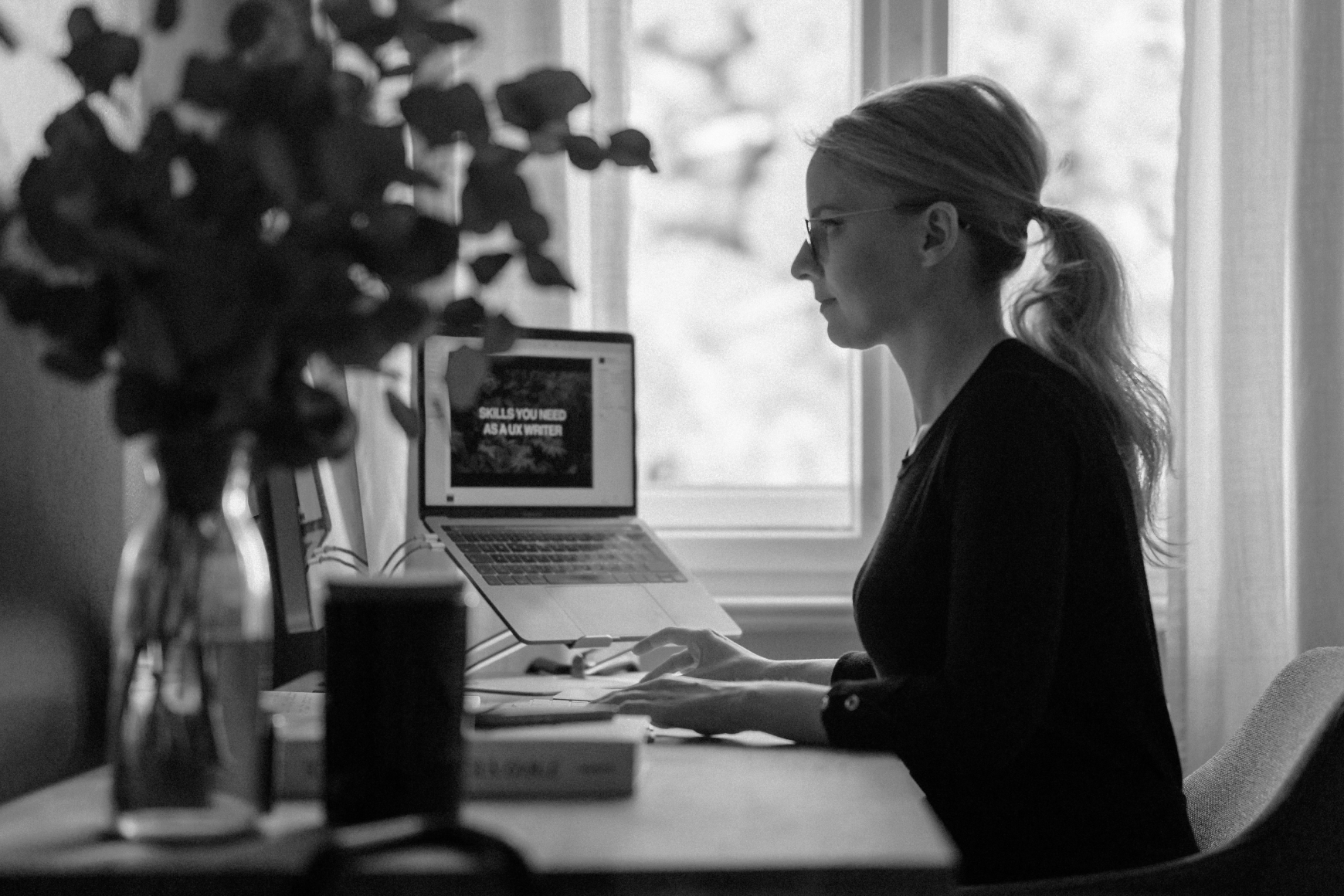 Dr Kat seen from the side, sitting at a desk, and using the keyboard of her laptop. The screen of the laptop is visible and shows a full screen image with a text written on it saying "Skills you need as a UX Writer"
