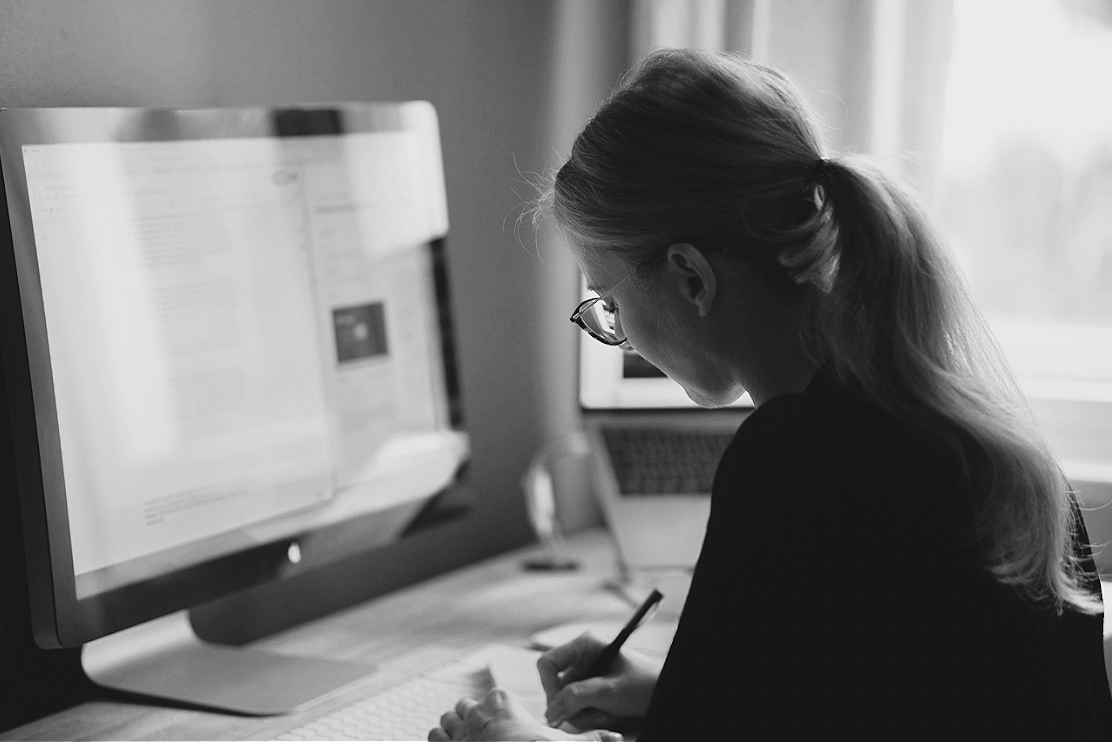 Dr Kat, sitting at her desk seen from behind, writing something down with a pen on paper. A laptop and display can be seen standing on the desk