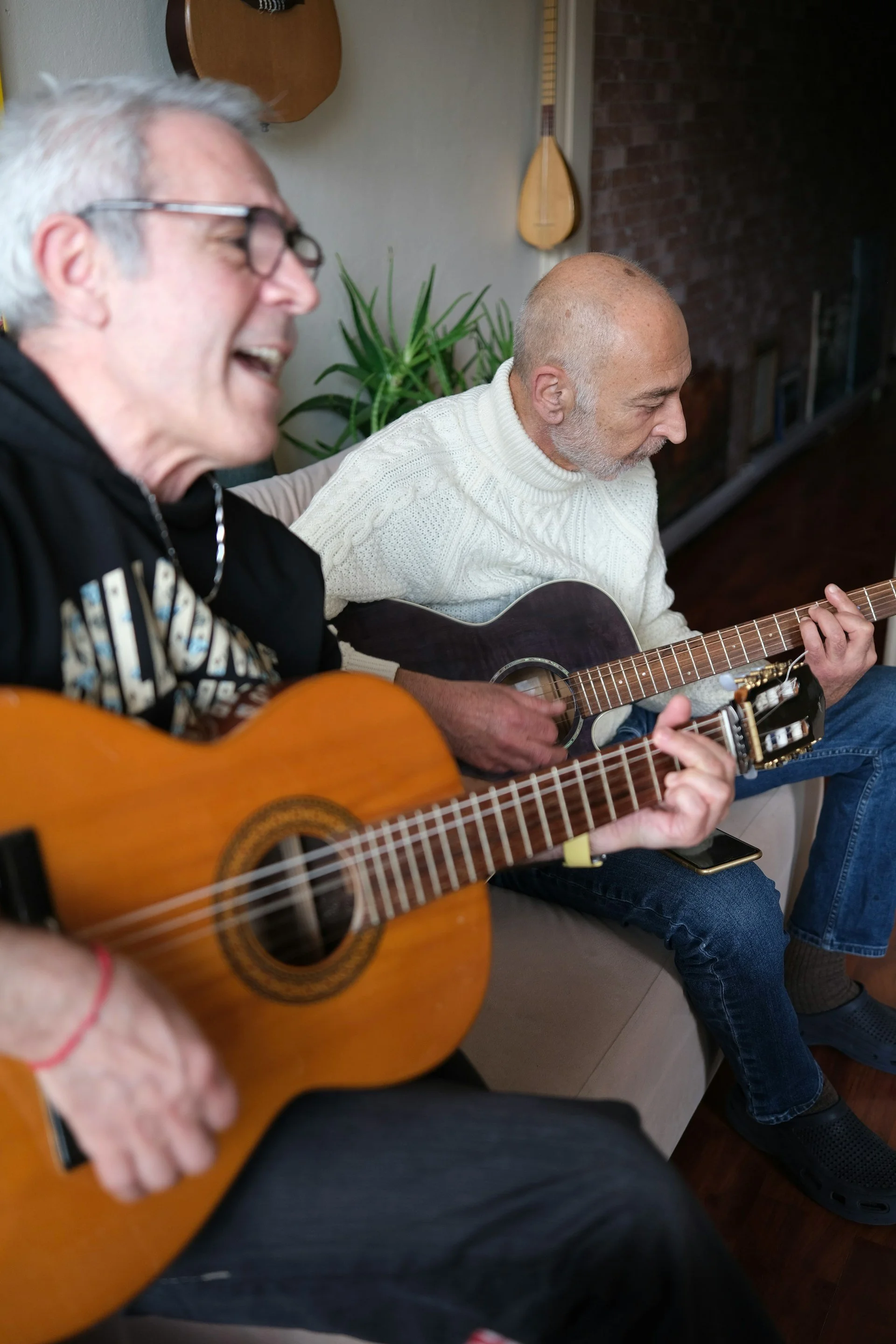 Two men play guitar together on a sofa