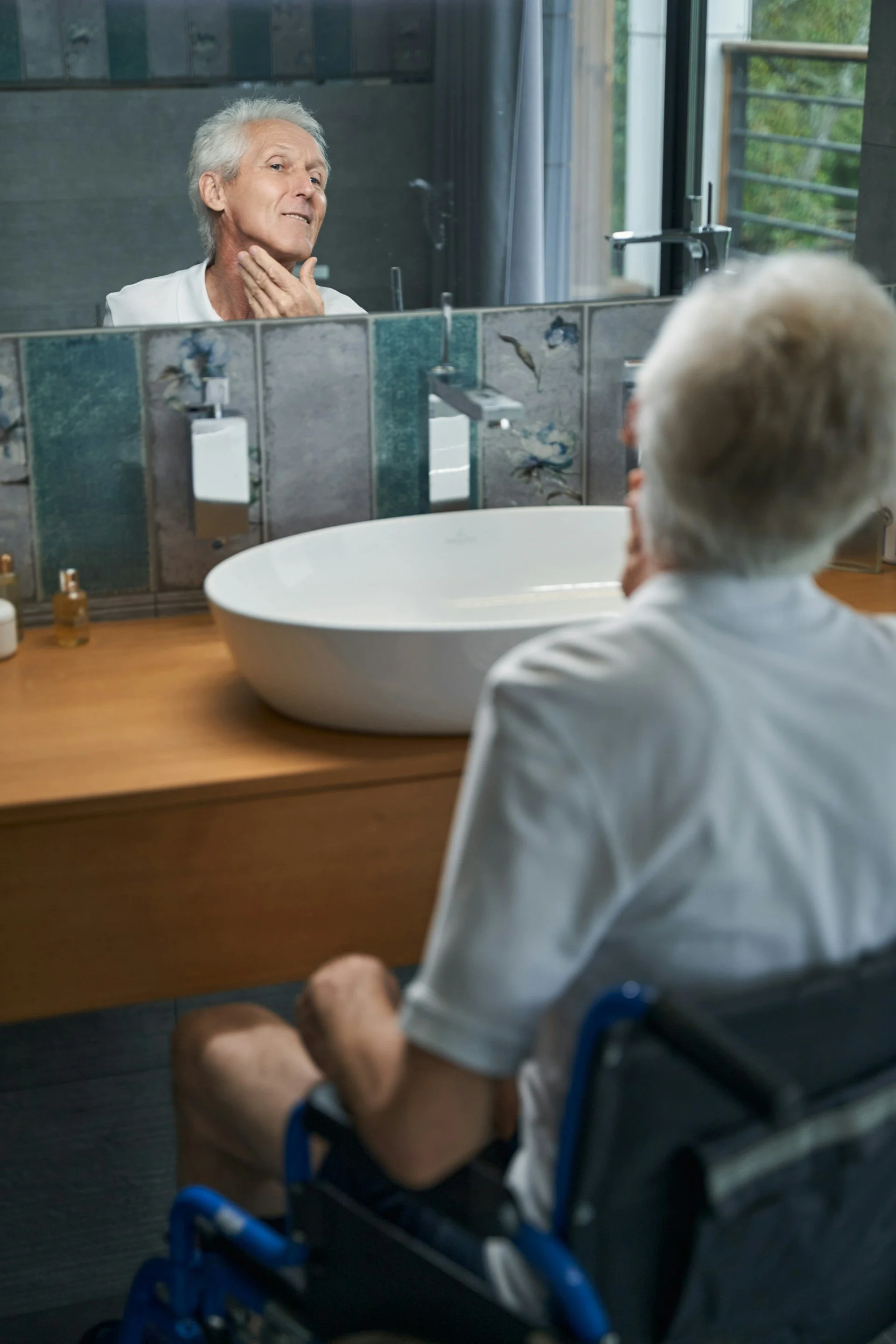 A man in a wheelchair inspects his face in his bathroom mirror