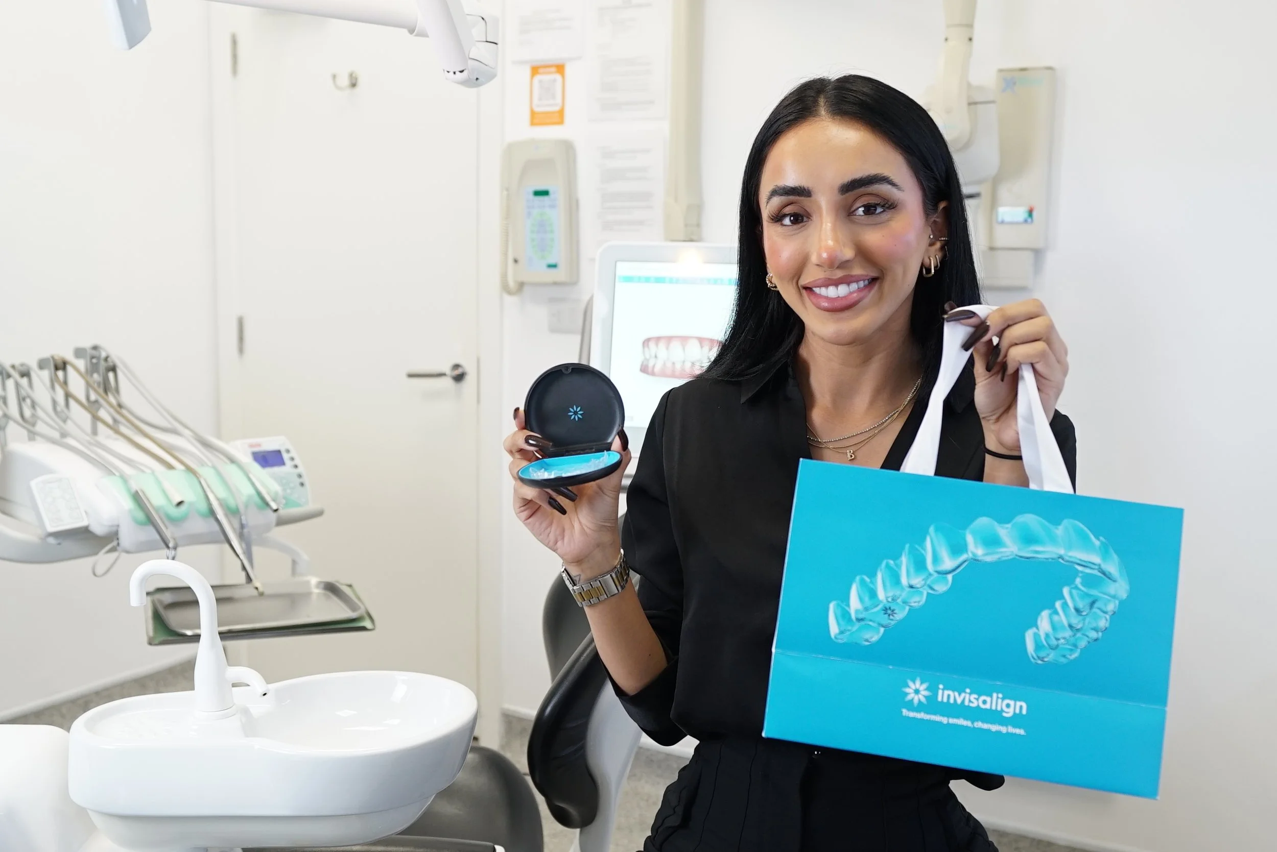 A woman smiling in a dental office, holding an Invisalign package and a cleaning case, with dental equipment and a teeth alignment image on a computer screen in the background.