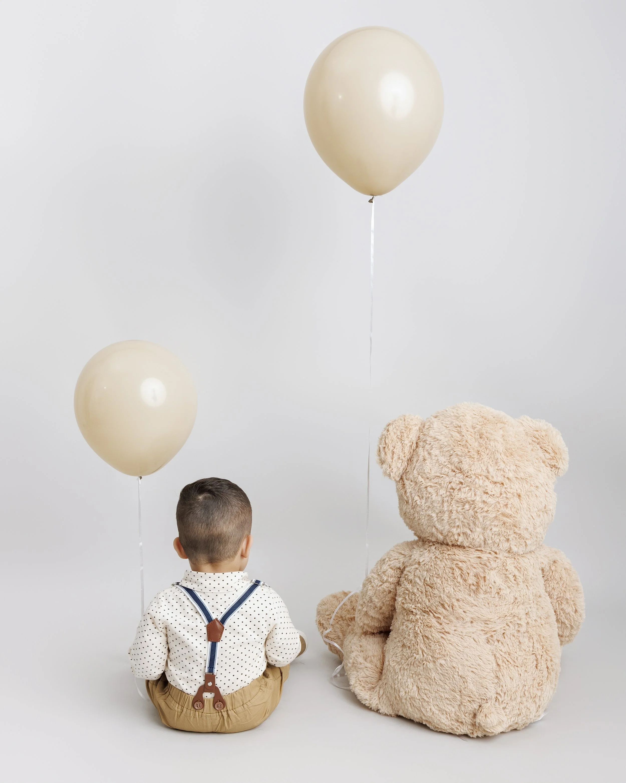 A young boy and a large teddy bear sitting on the floor with two beige balloons floating above them against a plain white background.