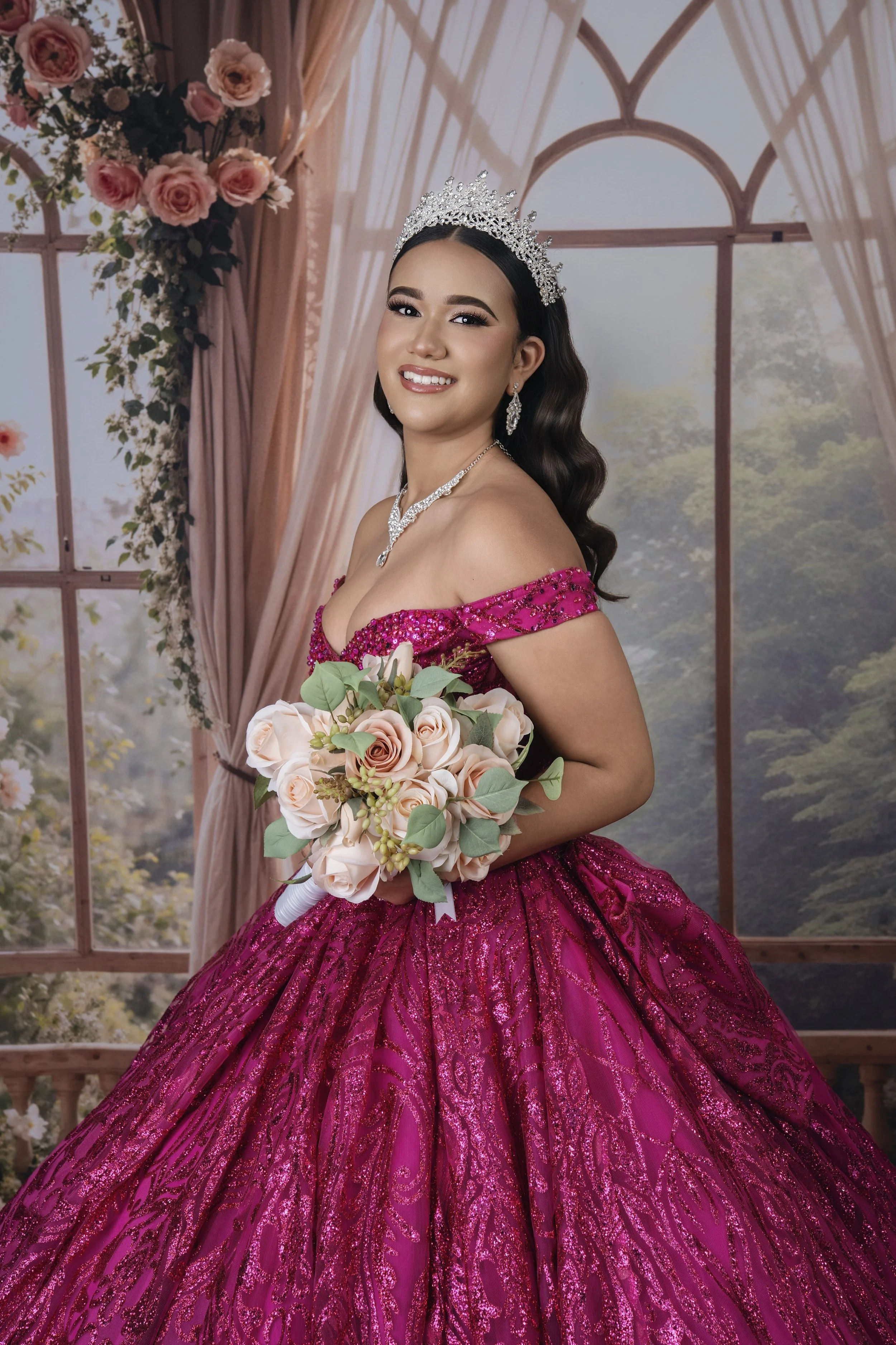 A woman dressed in a pink ball gown with sequins, wearing a silver tiara, holding a bouquet of pink roses and greenery, standing in front of a floral and curtain backdrop with trees visible outside.
