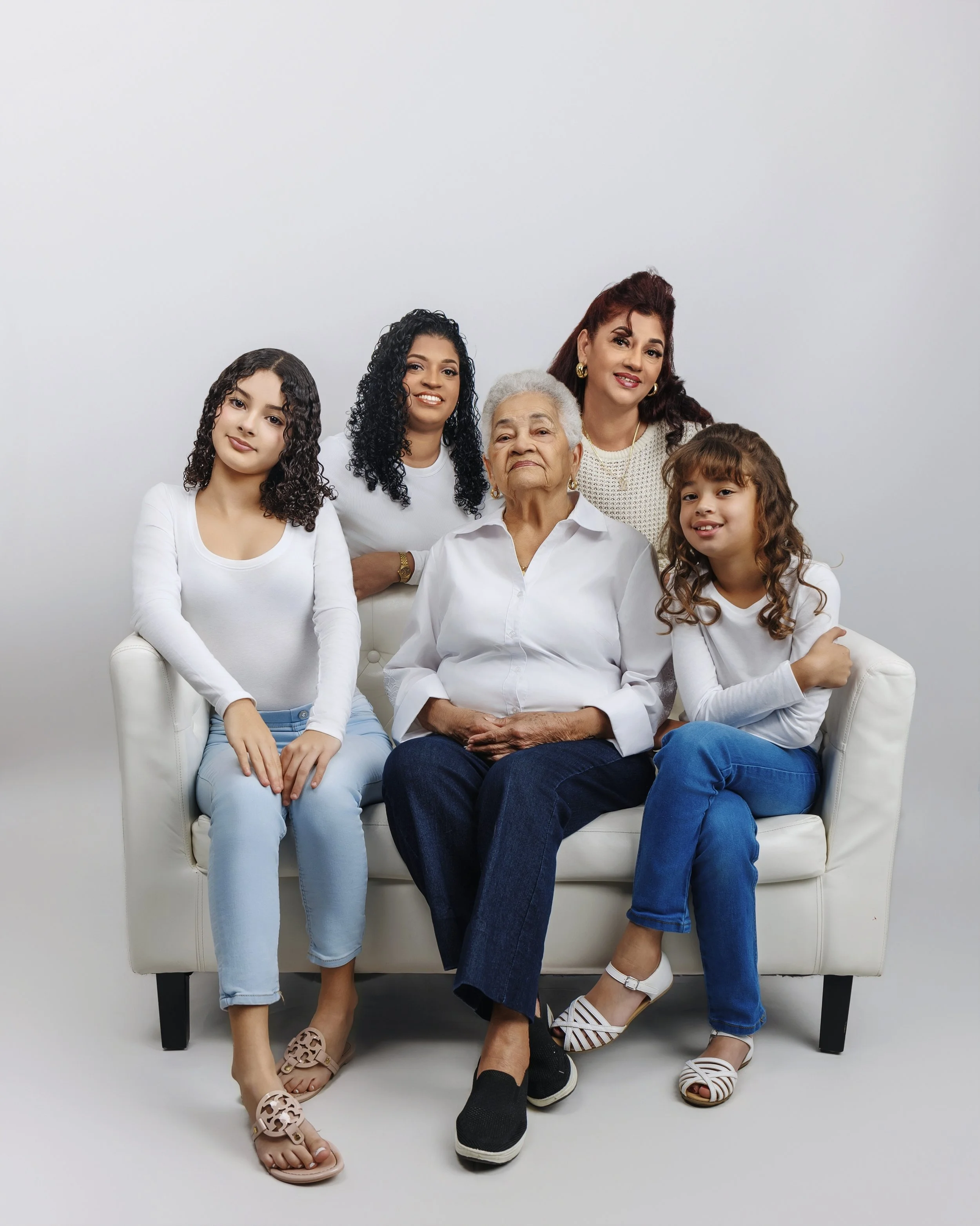 Five women, three young girls and two older women, sitting and standing together on a white sofa against a plain white background, all smiling and dressed in white and blue clothing.