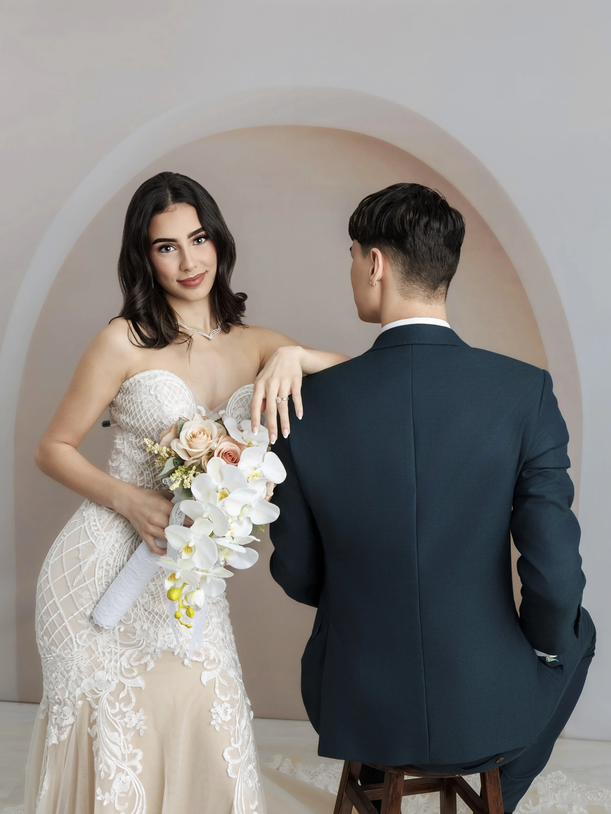 A woman in a white wedding dress holding a bouquet of roses and orchids, posing with a man in a dark suit sitting on a stool, facing her, in front of a beige arch backdrop.