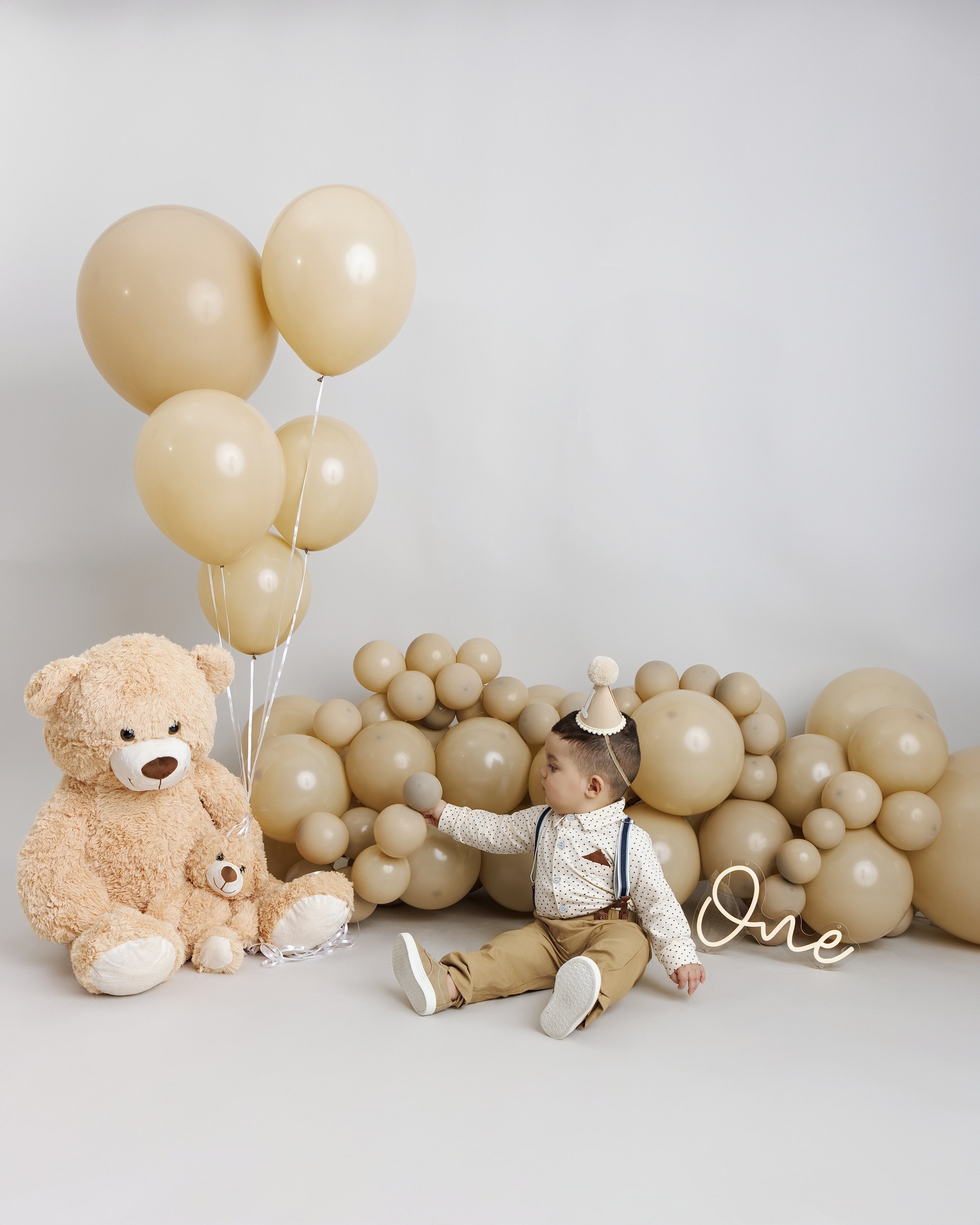 Young child sitting on floor with balloons, teddy bear, and decorative balloons spelling 'one' in neutral tones for first birthday celebration.