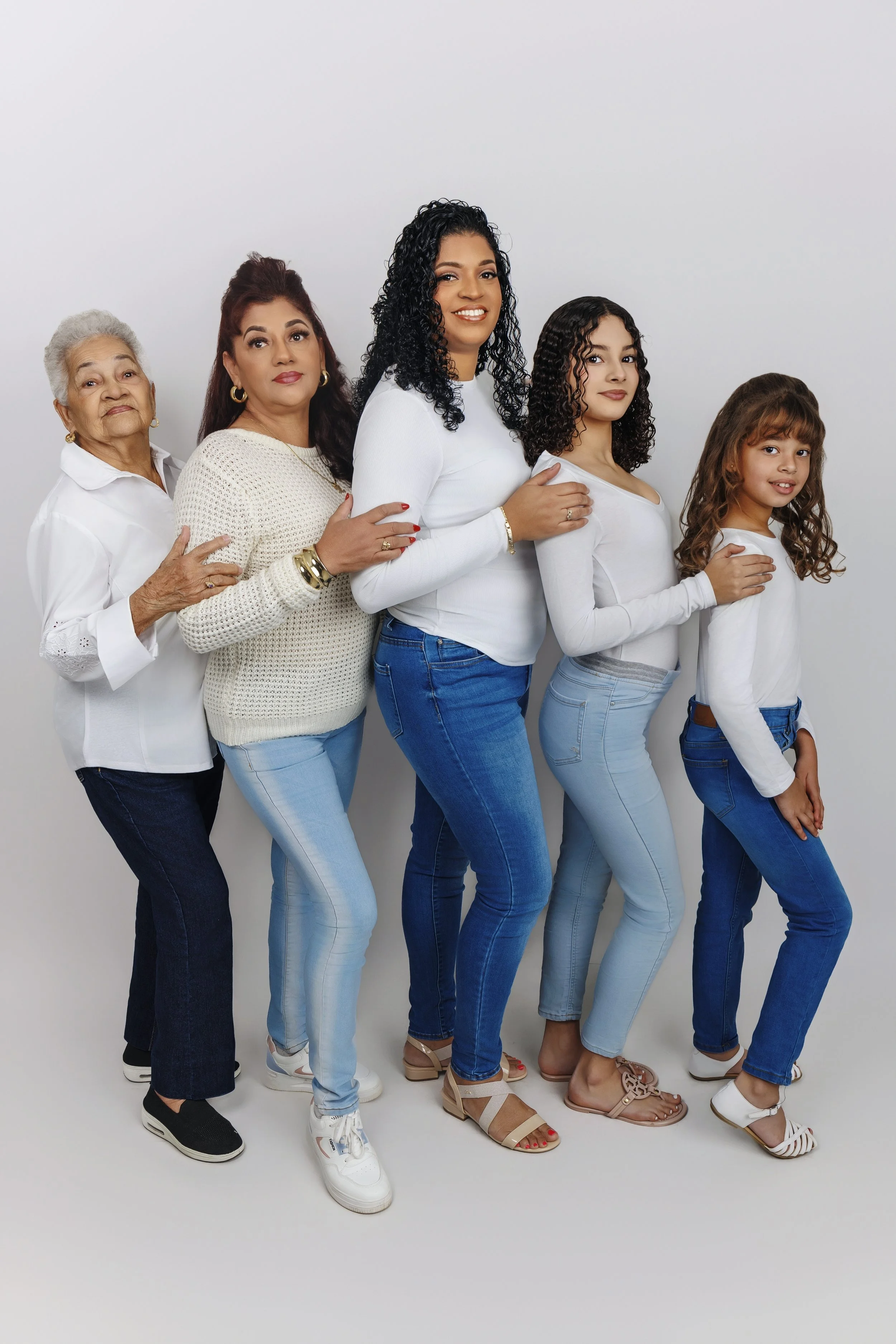 A multigenerational African American family poses together against a plain white background, standing in a line with hands on each other's shoulders, dressed in white tops and blue jeans.
