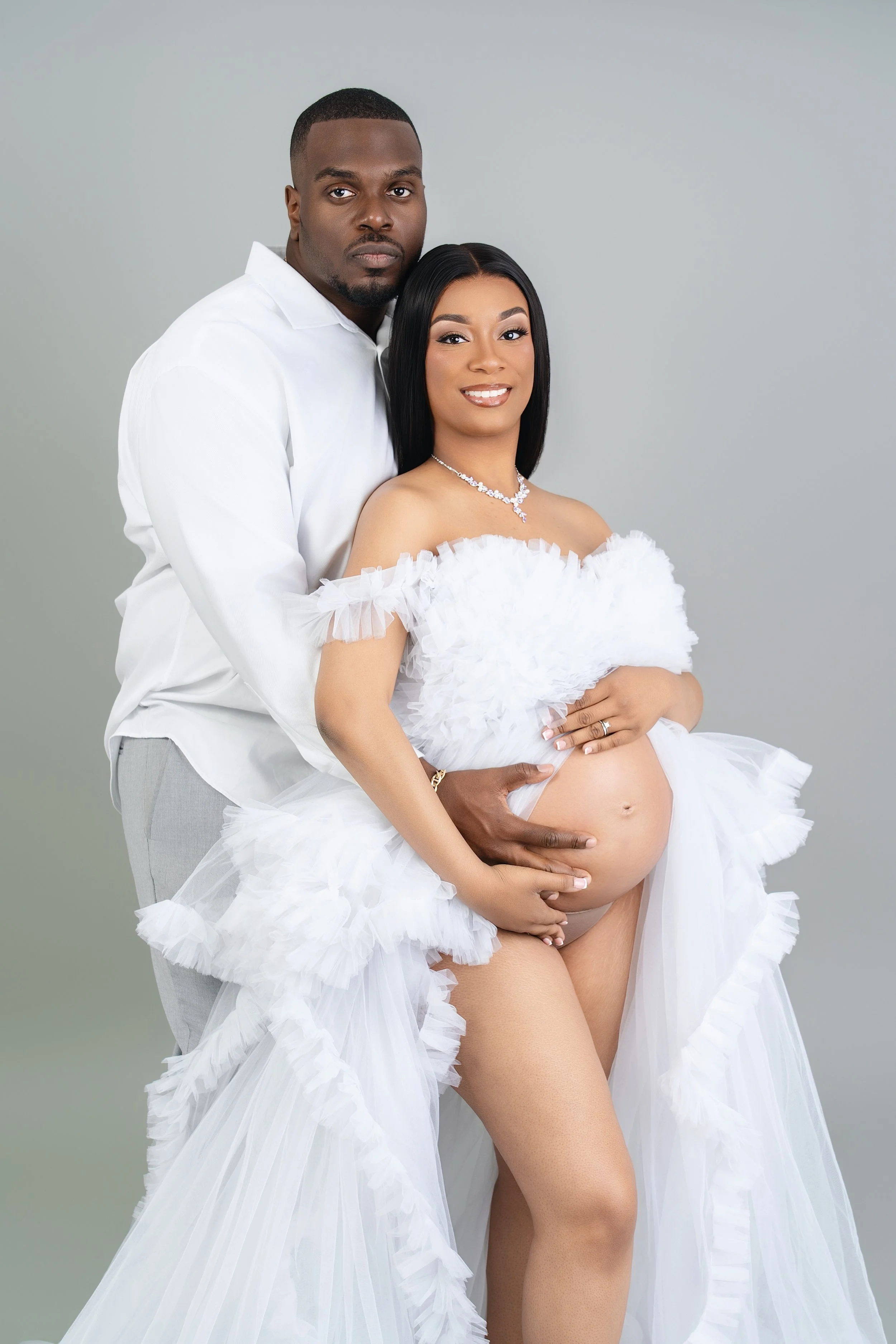 A pregnant woman in a white, ruffled dress with a strapless top, standing with her partner. The man is in a white shirt and light gray pants, standing behind her with his arms around her, both smiling. The woman is wearing jewelry including a necklace and earrings, with her hand on her belly and her partner's hand on her waist.