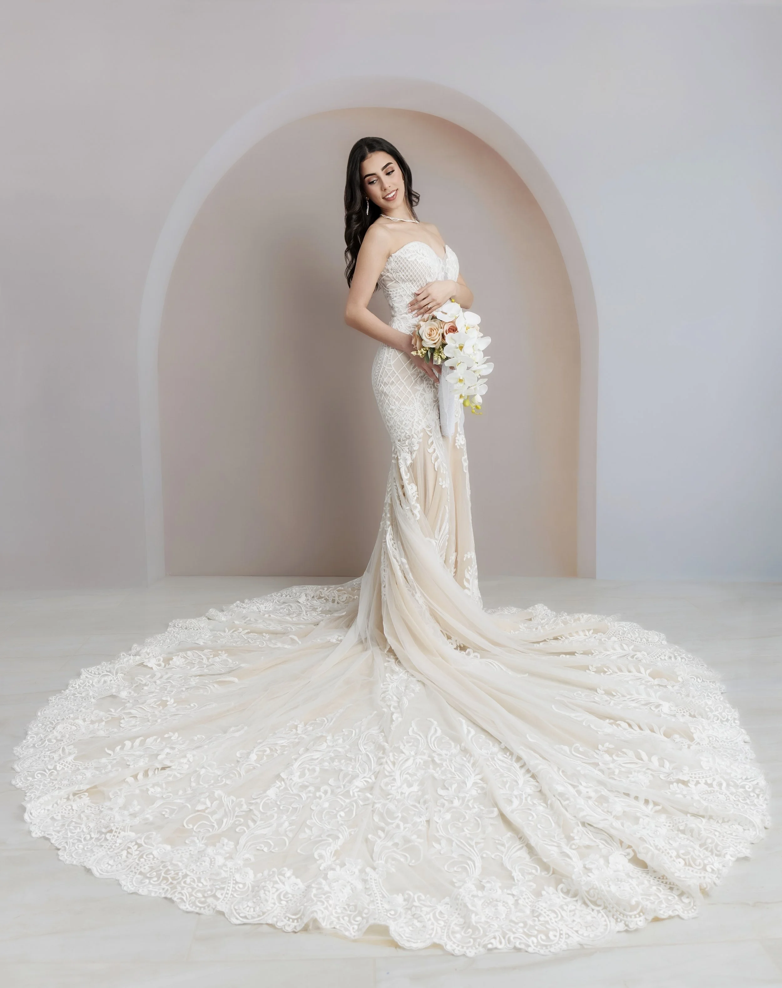 Bride in a white wedding dress holding a bouquet of roses and orchids, standing in front of a neutral-colored wall with an arched niche.