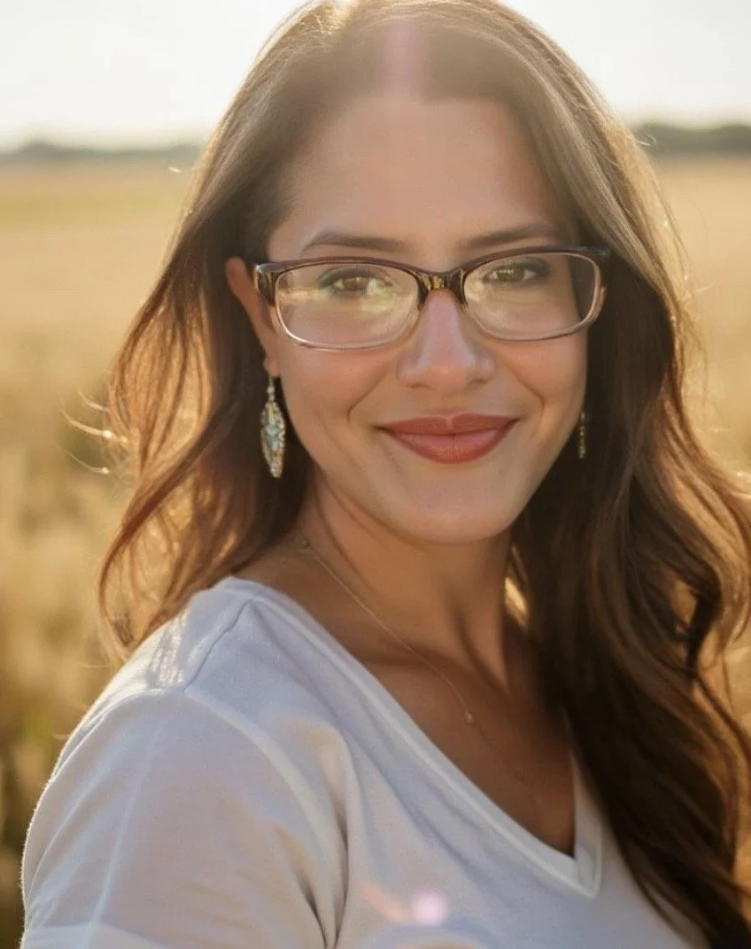 A smiling woman with glasses and earrings in a white shirt outdoors during sunset.