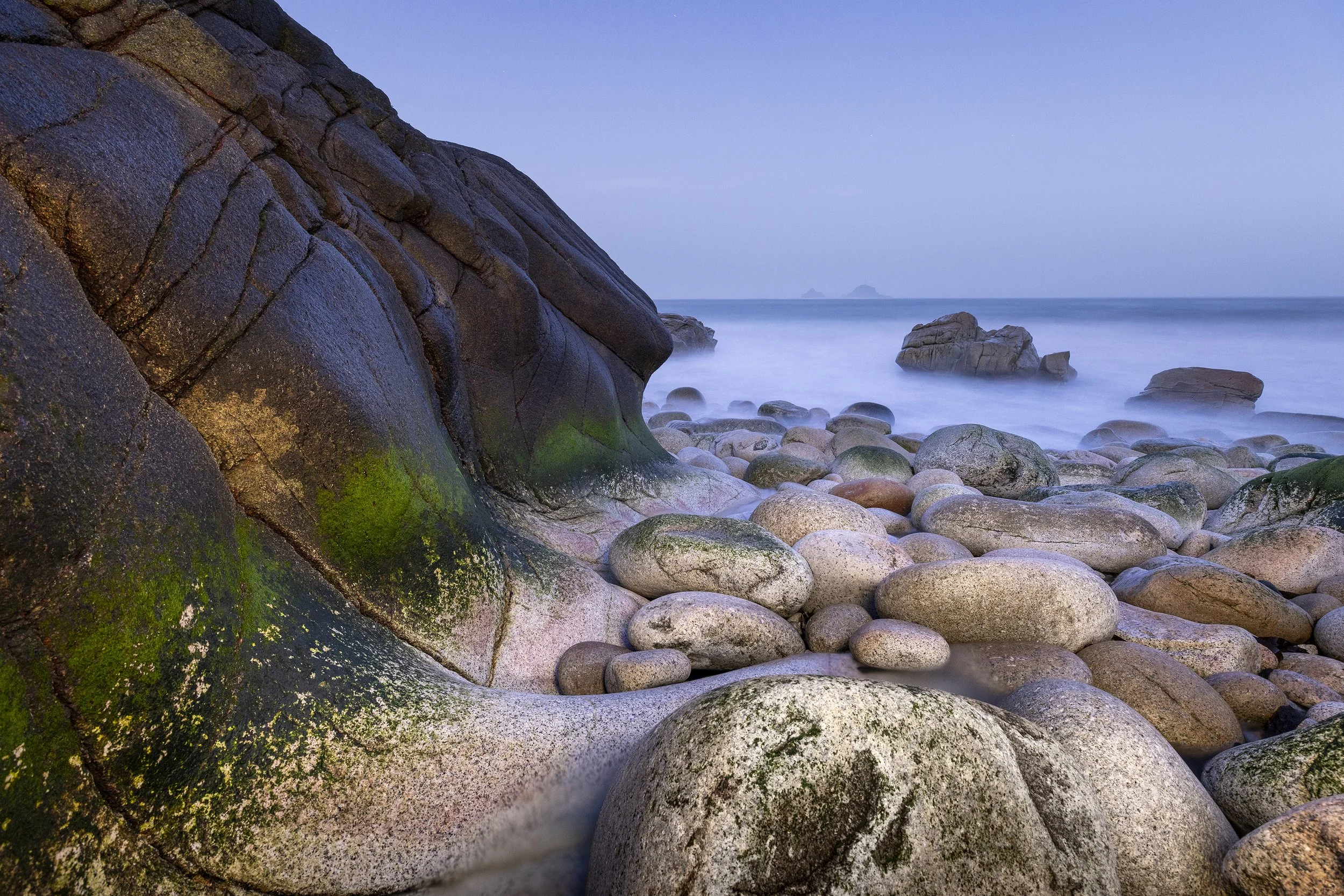 Rocky shoreline with large moss-covered rocks and a calm ocean in the background at dusk or dawn.