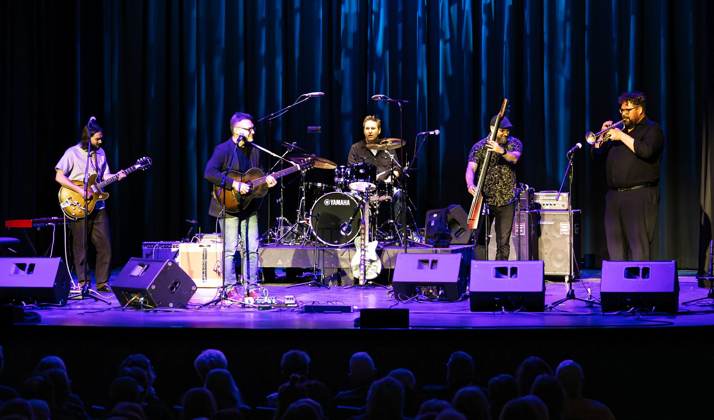 A band performing on stage with five musicians playing instruments, including guitars, bass, drums, and trumpet, in front of an audience.